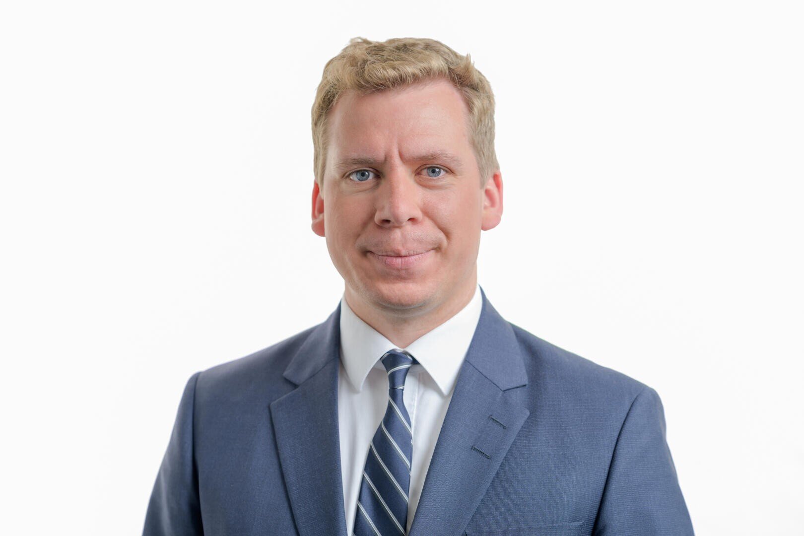 A man with short blond hair, wearing a blue suit, a white shirt and a striped tie, stands in front of a plain white background and smiles slightly.