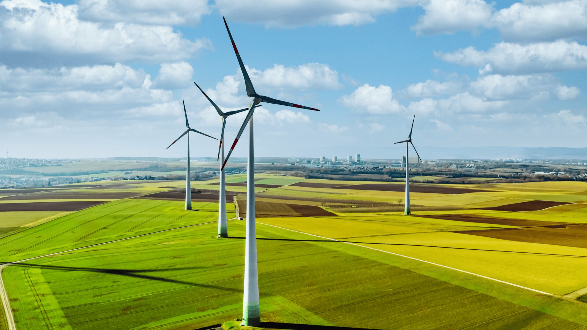 Four wind turbines stand in a patchwork of green and yellow fields under a partly cloudy sky, generating renewable energy in a rural landscape. Distant buildings are visible on the horizon.