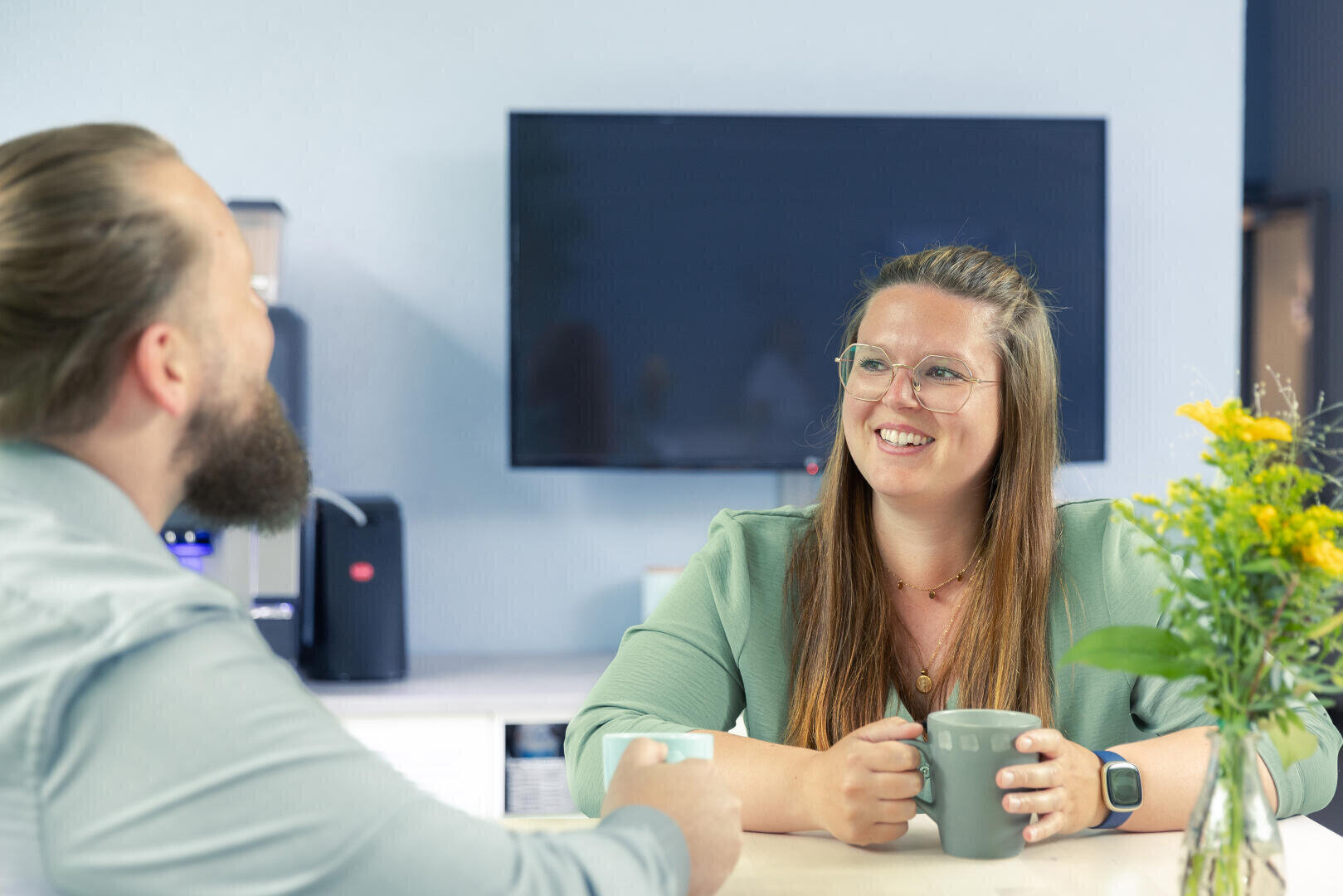 A woman and a man sit at a table, smiling and talking while holding mugs. There is a vase with yellow flowers on the table, and a large television screen in the background.