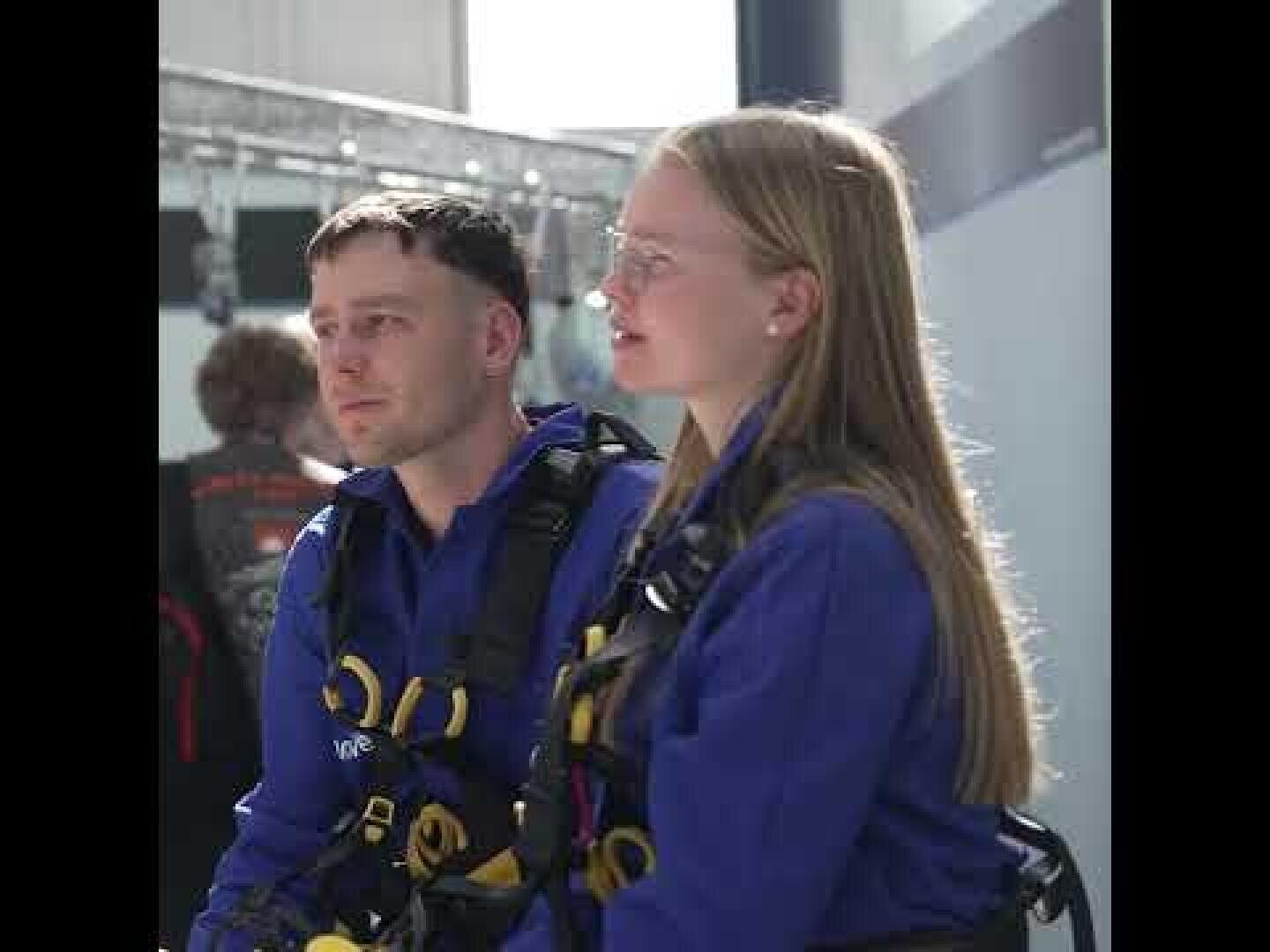 Zwei Personen in blauen Uniformen und Klettergurten sitzen in einem Innenraum nebeneinander und schauen nach vorne. Der Hintergrund ist unscharf, eine weitere Person und einige Geräte sind zu sehen.