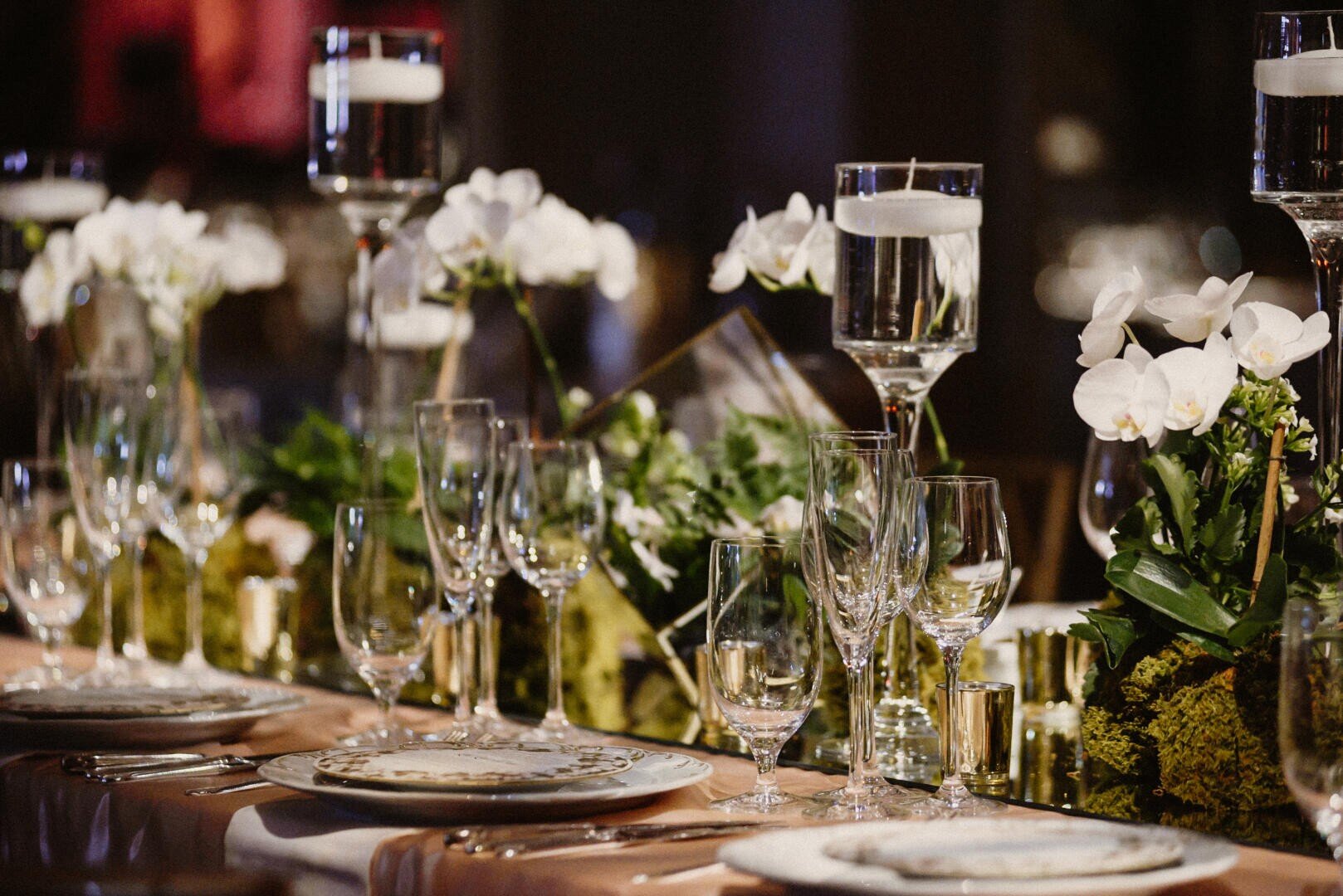 Elegant table decoration with white flowers, glass candle holders with floating candles and several glasses on a white tablecloth for a formal event or celebration.
