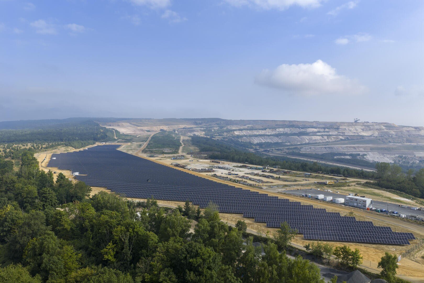 A large solar park with rows of solar panels is located near a forest, with industrial buildings, open land and distant hills under a partly cloudy sky.