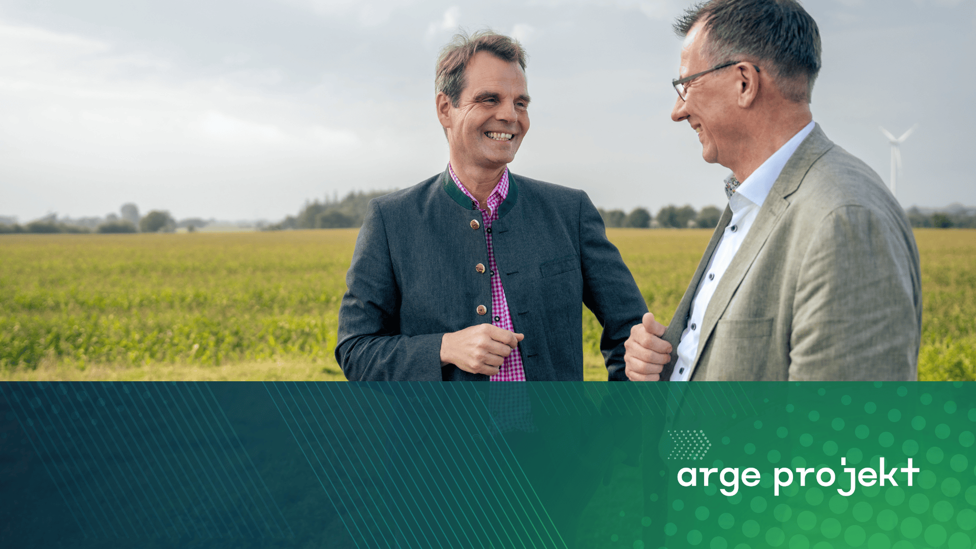 Zwei Männer in Jacken stehen lächelnd und redend auf einer grünen Wiese mit einer Windkraftanlage im Hintergrund. Im Vordergrund befindet sich eine blaue und grüne Überlagerung mit dem Text "arge projekt.".