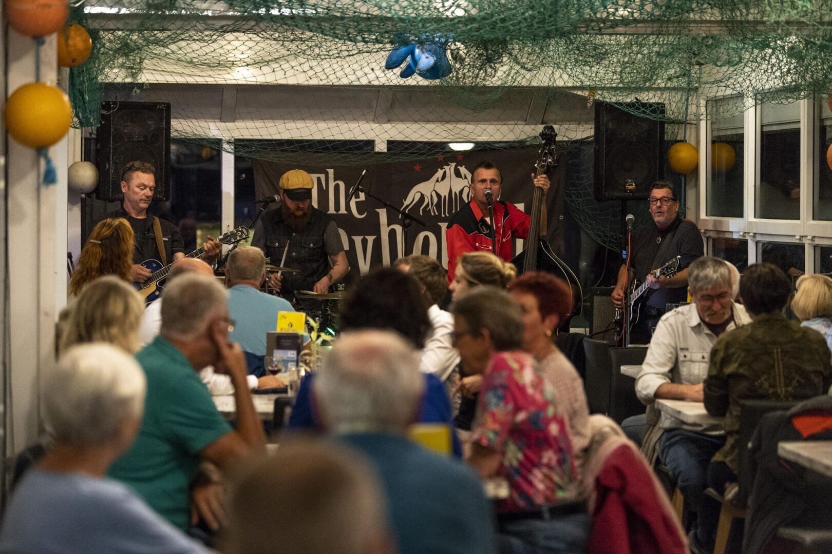 A band plays live music on a small indoor stage decorated with fishing nets, while a crowd of seated people watch and dine at tables in a casual, bustling restaurant atmosphere.