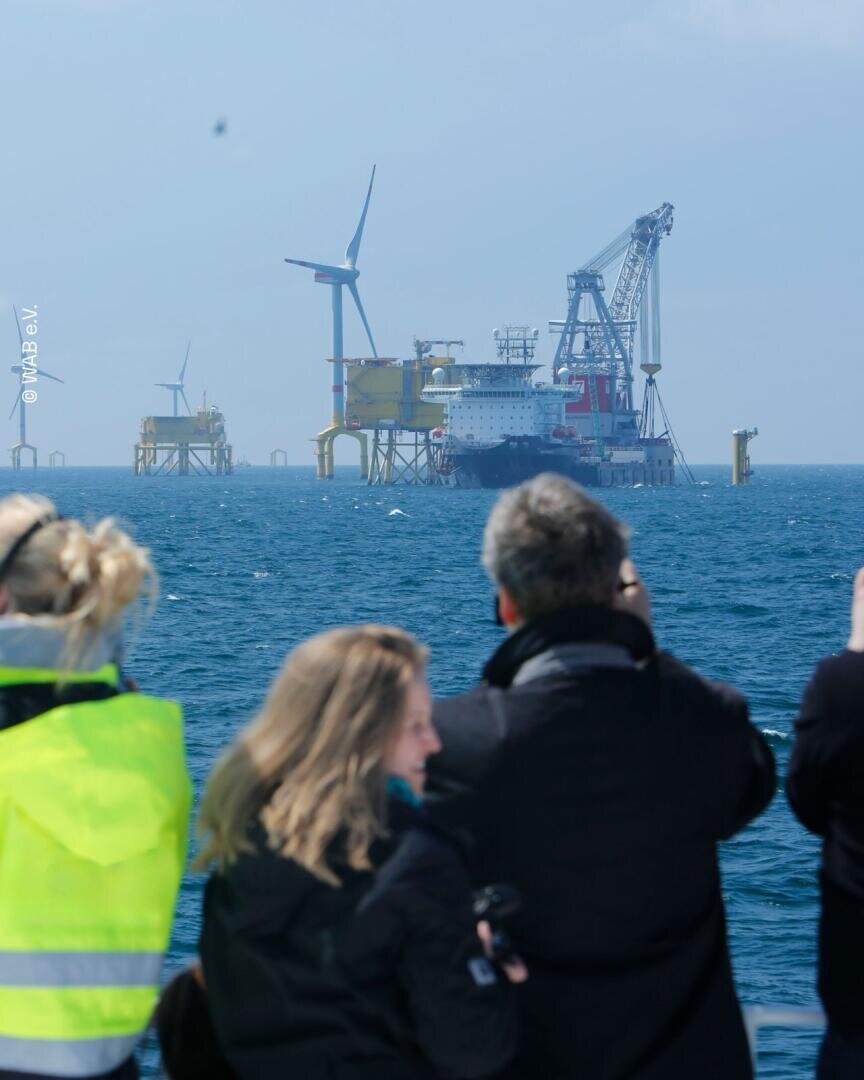 Mehrere Personen betrachten Offshore-Windkraftanlagen und Industrieplattformen auf dem Meer, wobei unter dem bewölkten Himmel Kräne und Ausrüstung zu sehen sind.