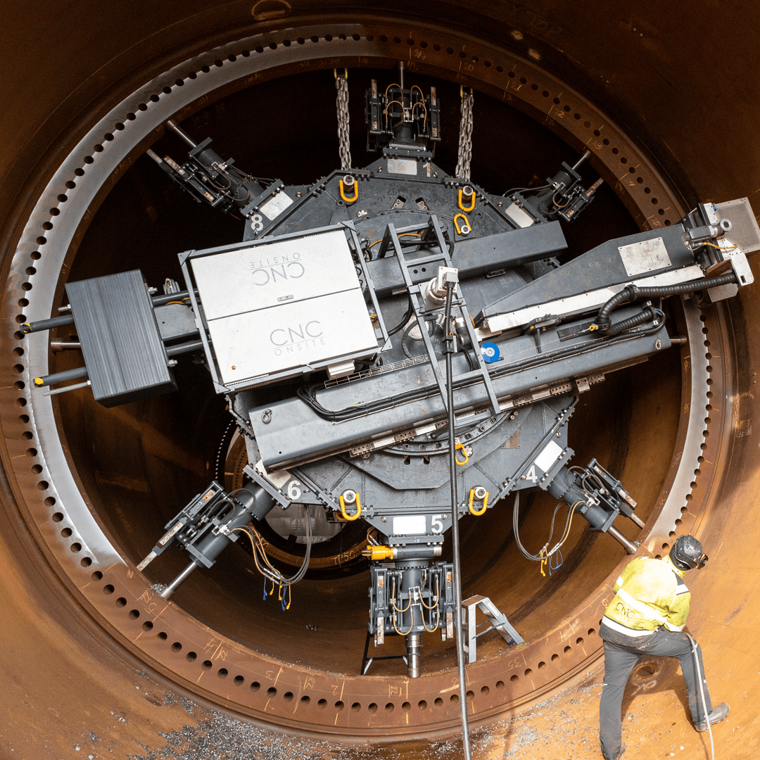 A worker in a yellow safety jacket and helmet stands inside a large cylindrical metal structure, operating heavy industrial machinery attached to the interior wall.