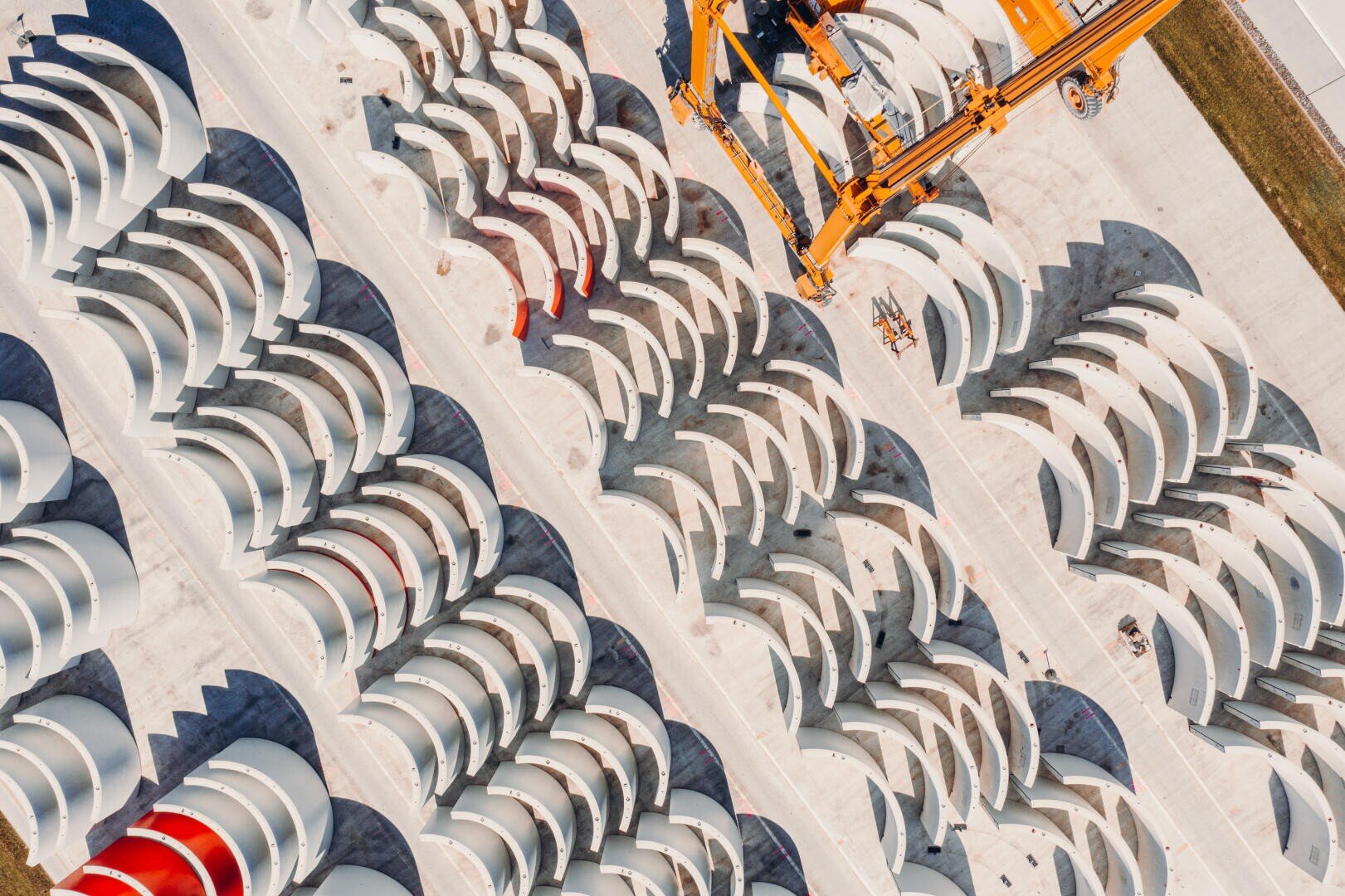 Luftaufnahme von fein säuberlich angeordneten Reihen großer weißer Windturbinenflügel auf einer Betonfläche, mit orangefarbenen Kränen in der Nähe, die Schatten auf die Szene werfen.