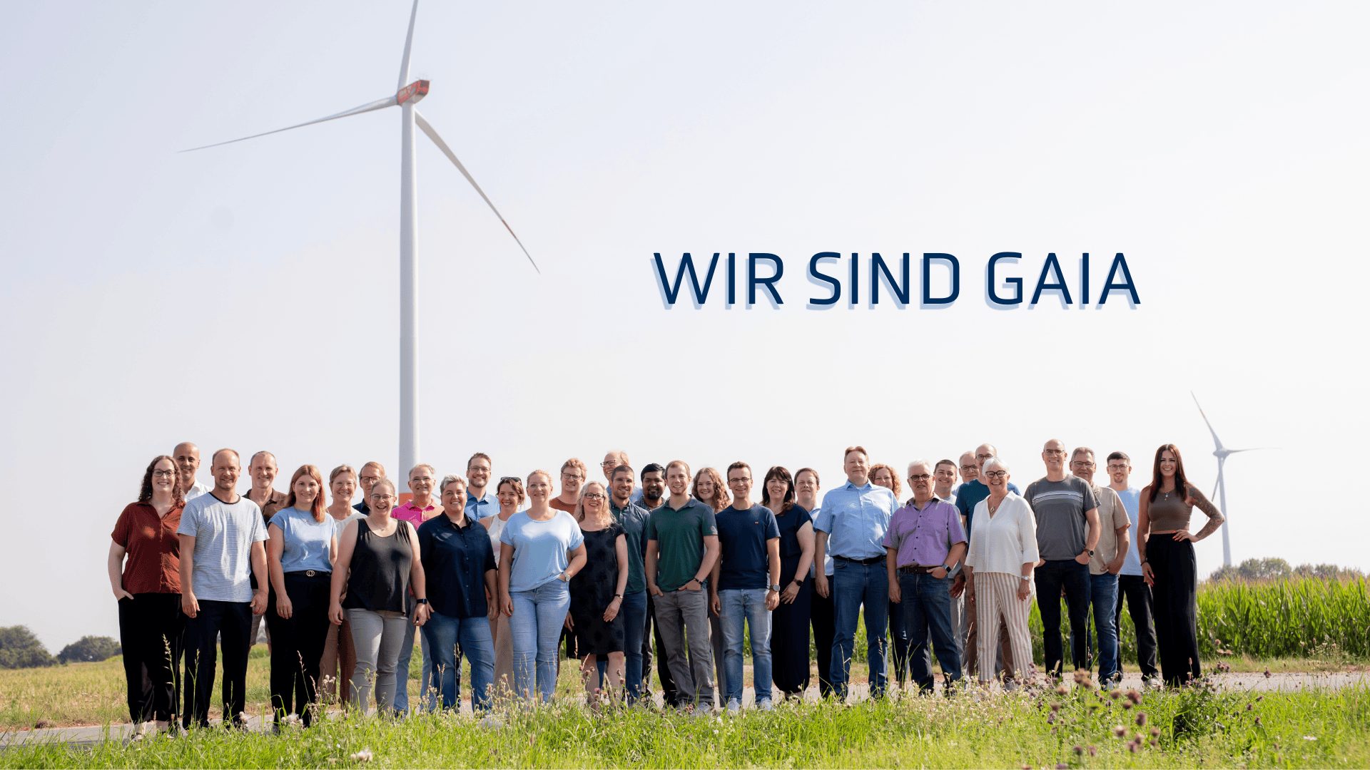 Eine Gruppe von etwa 35 Personen steht im Freien vor Windrädern und einem Feld zusammen. Über ihnen prangt in großen Lettern der Text WIR SIND GAIA.