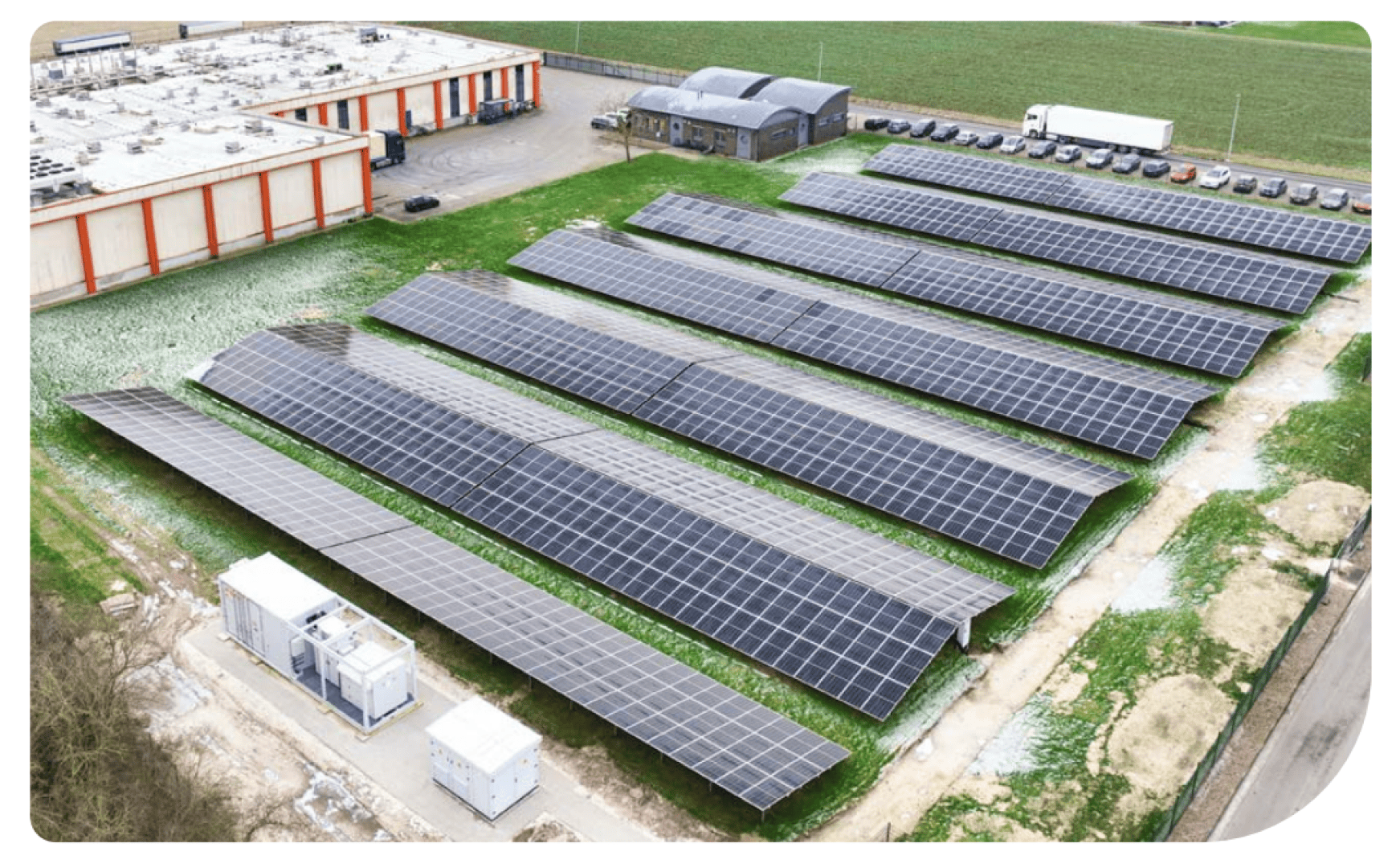 Luftaufnahme von mehreren Reihen von Solarmodulen, die auf einer Wiese in der Nähe mehrerer Gebäude und geparkter Fahrzeuge installiert sind, mit Feldern im Hintergrund.