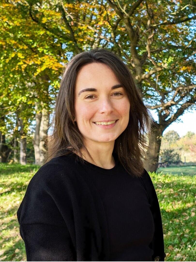 A woman with shoulder-length brown hair and a black top smiles outdoors, standing in a sunlit park with green grass and leafy trees in the background.
