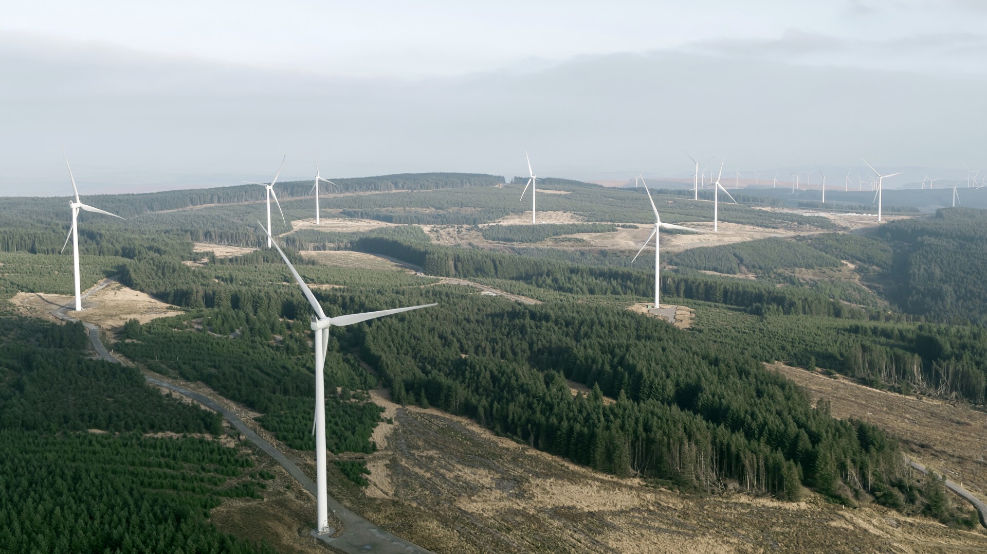 Aerial view of a wind farm with multiple large white wind turbines scattered across a hilly, forested landscape under a cloudy sky.