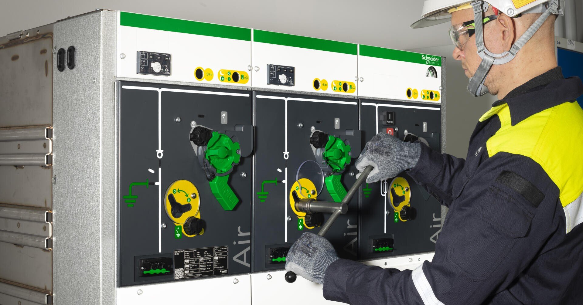 Ein Techniker mit Schutzausrüstung betätigt die Schalter einer elektrischen Schalttafel mit der Aufschrift Air von Schneider Electric. Die Schalttafel ist mit farbigen Bedienelementen, Messinstrumenten und Kontrollleuchten ausgestattet.