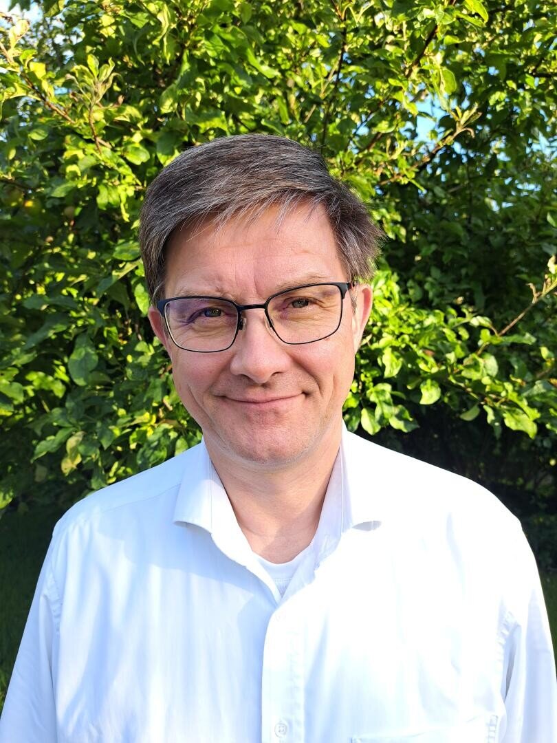 Ein Mann mittleren Alters mit kurzen braunen Haaren und Brille, der ein weißes Hemd mit Kragen trägt, steht im Freien vor grünen Bäumen und lächelt im hellen Sonnenlicht in die Kamera.