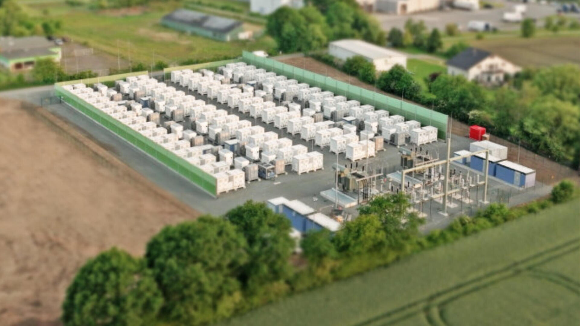 Luftaufnahme einer eingezäunten Anlage mit Reihen von weißen Containern, wahrscheinlich Batteriespeicher und elektrischer Infrastruktur, umgeben von Bäumen und nahe gelegenen Gebäuden.