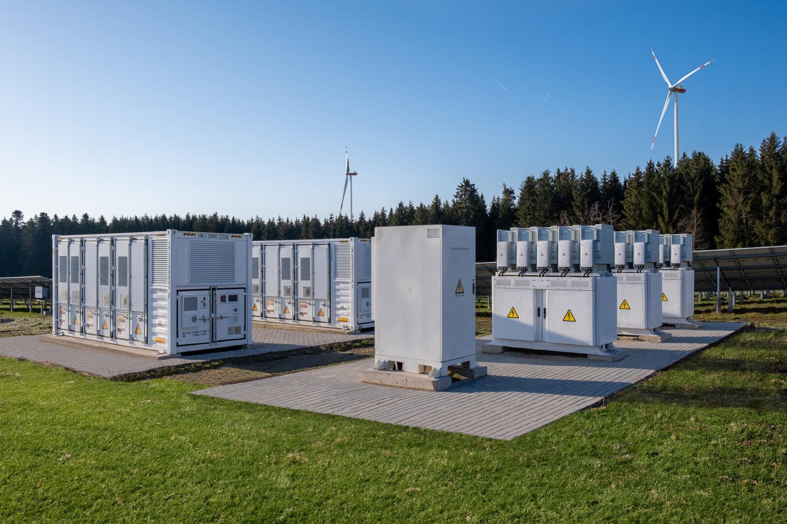 Ein Speicher für erneuerbare Energien mit weißen Batteriecontainern und elektrischen Geräten auf einer Wiese, rechts Sonnenkollektoren und im Hintergrund Windturbinen und Bäume unter einem klaren blauen Himmel.