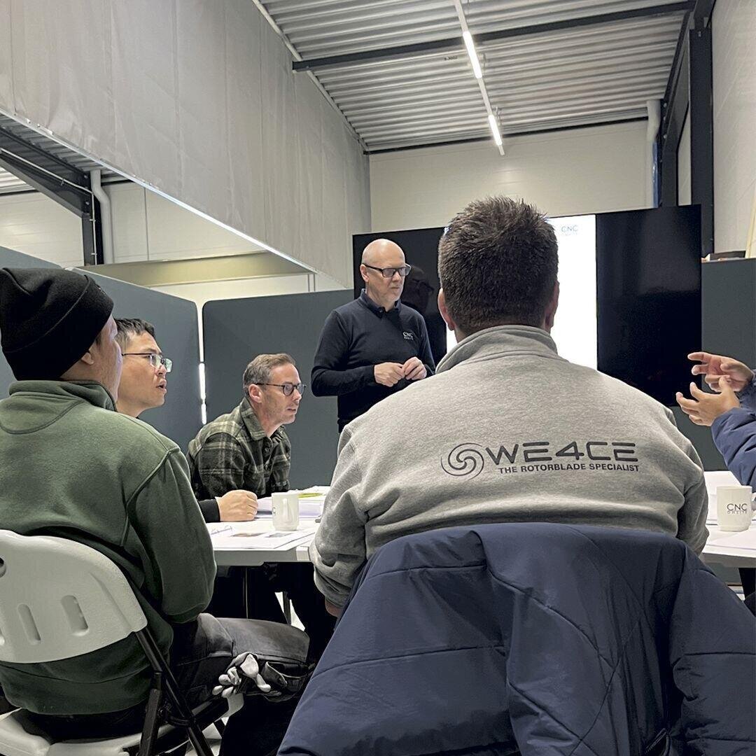 A group of people sits around a table in a meeting room, listening to a man standing and speaking. One attendee wears a jacket with “WE4CE The Rotorblade Specialist” on the back. Papers and cups are on the table.