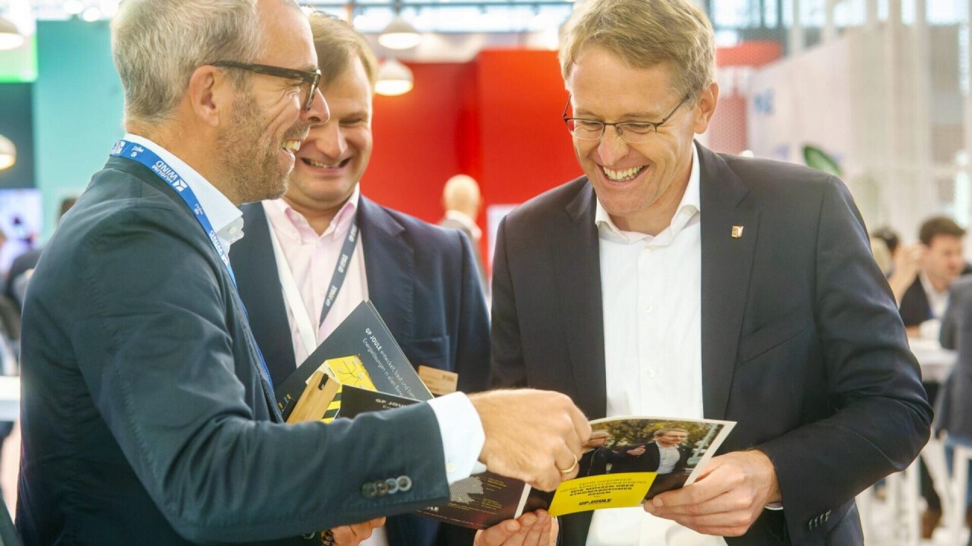 Three men in business attire stand together smiling and looking at brochures in a brightly lit indoor setting, possibly at a professional event or conference.