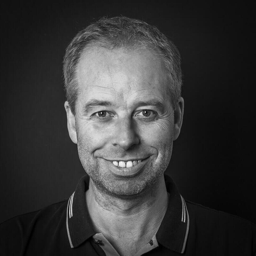 Black and white portrait of a smiling middle-aged man with short, light-colored hair, wearing a dark polo shirt with a light hem, against a plain dark background.