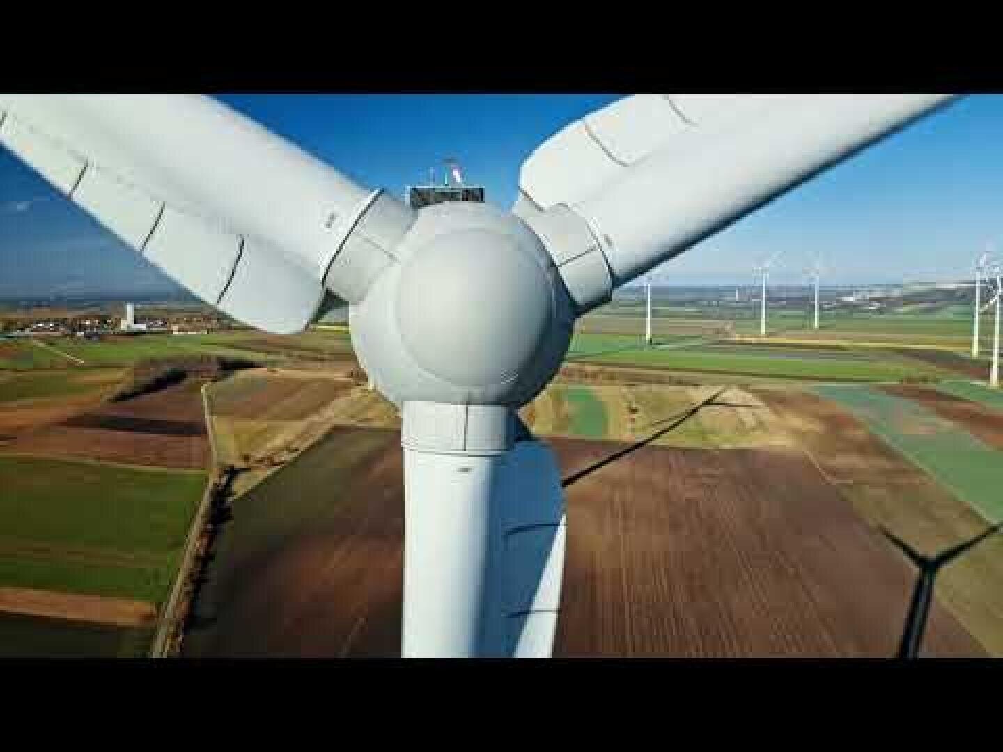 Close-up view of the top of a wind turbine with its blades, set above patchwork fields and several other wind turbines in the background under a clear blue sky.