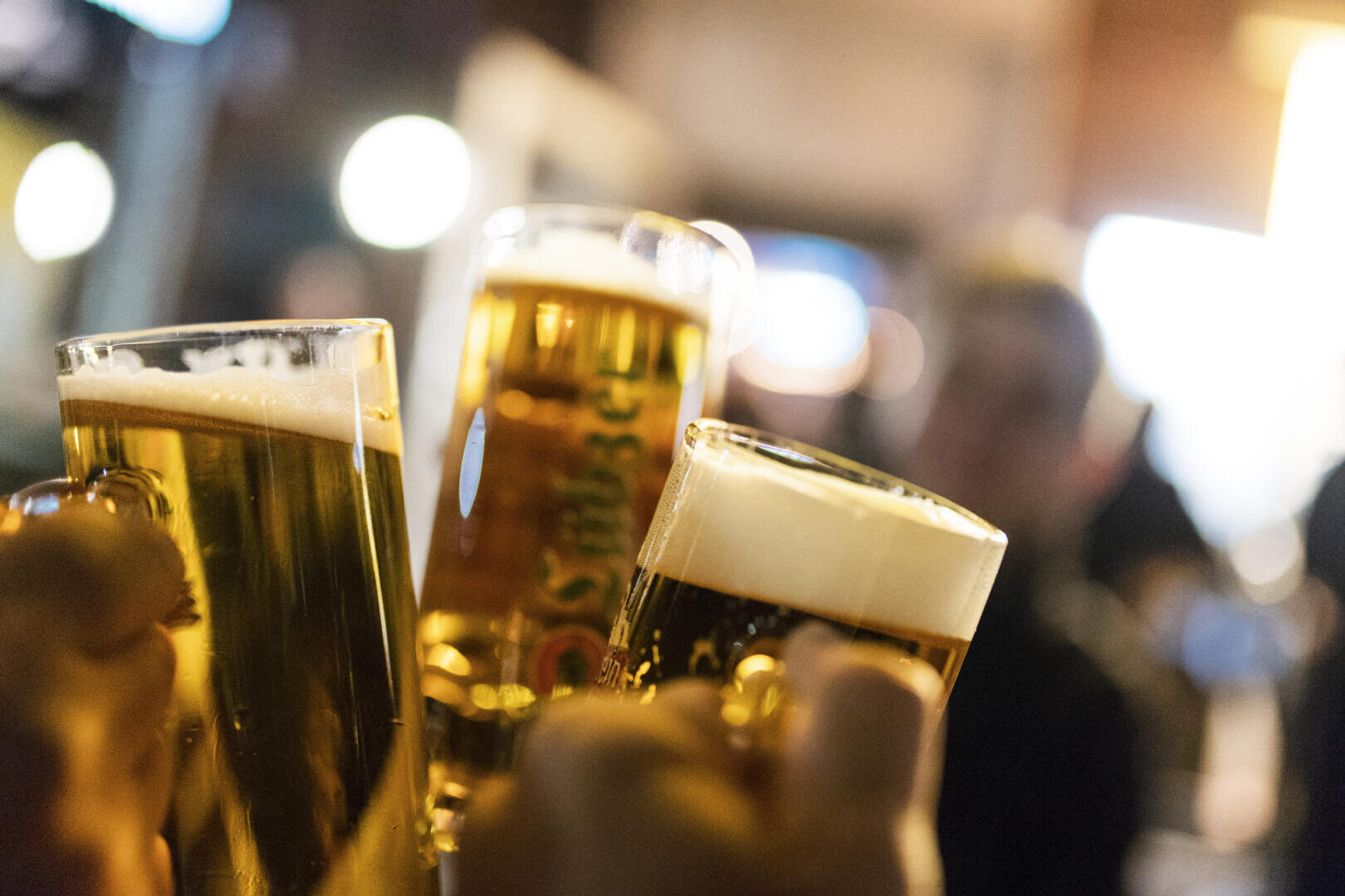 Three hands hold beer glasses and clink glasses. In the background, people and warm lights are blurred, suggesting a lively gathering or celebration.