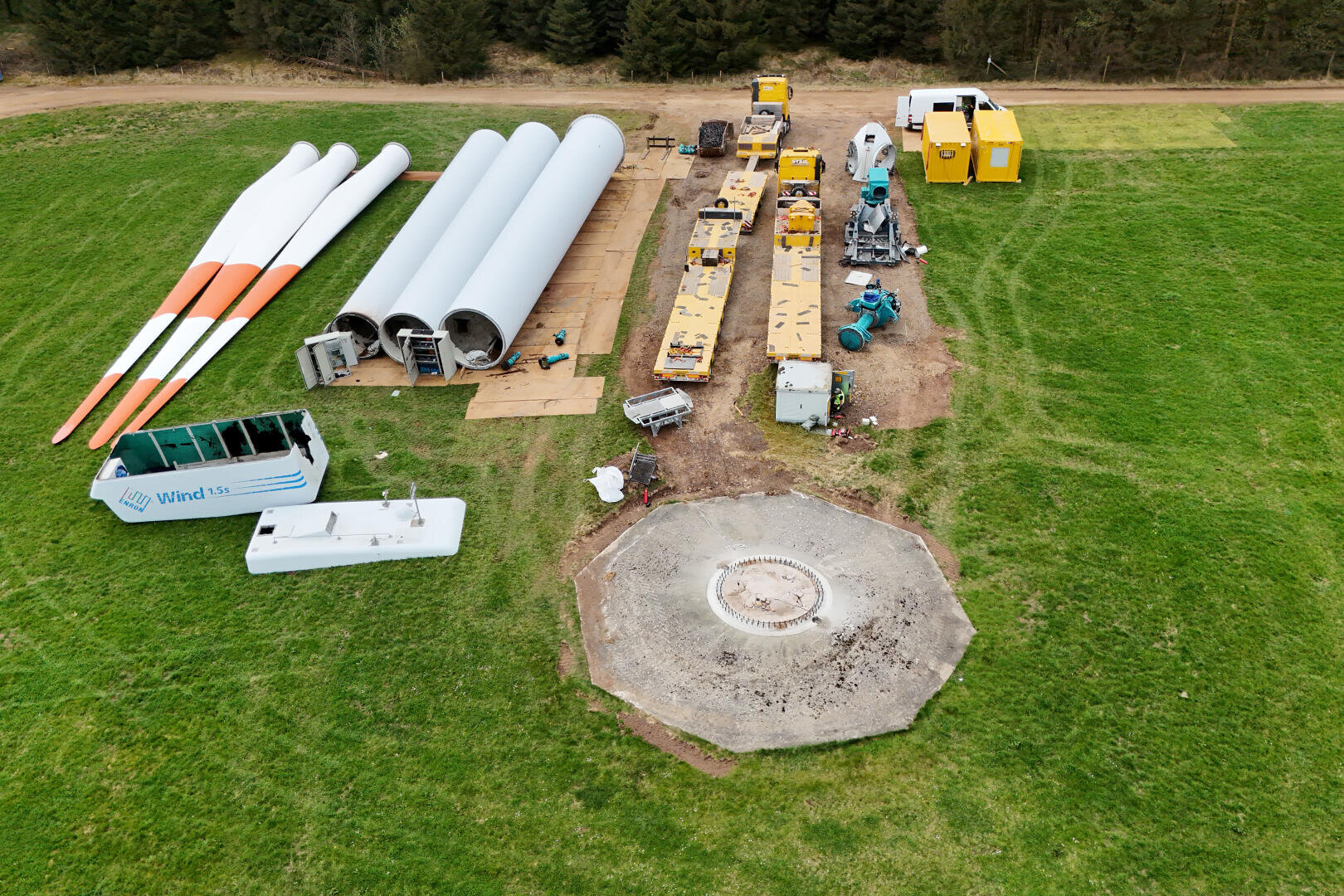 Luftaufnahme von Windturbinenteilen, Baufahrzeugen und Ausrüstungen, die auf einer Wiese in der Nähe eines runden Betonfundaments angeordnet sind.