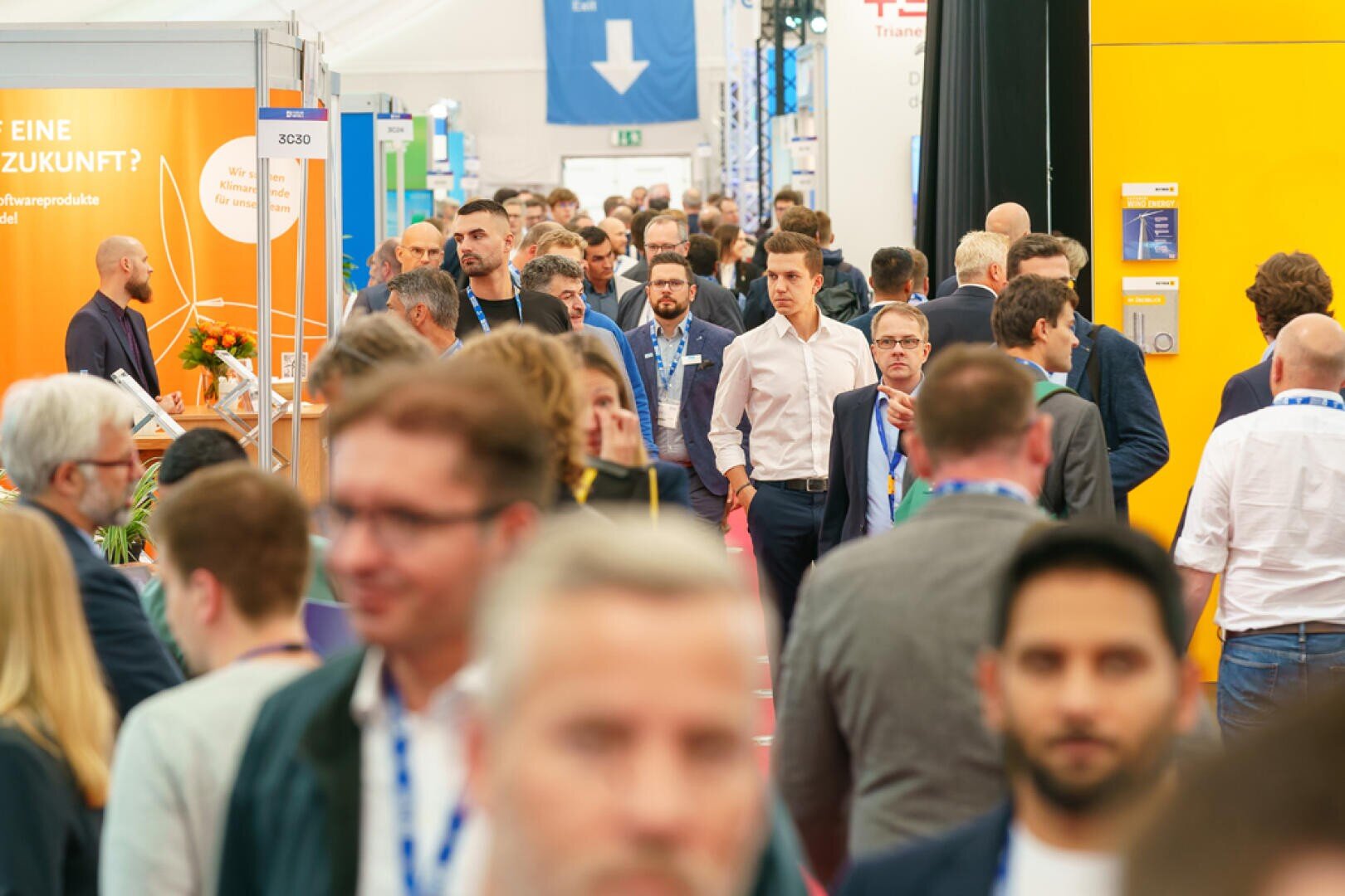 A crowded exhibition or conference hallway with lots of people walking and talking, surrounded by colorful booths and displays on both sides. Most of those present are wearing business attire and name badges.