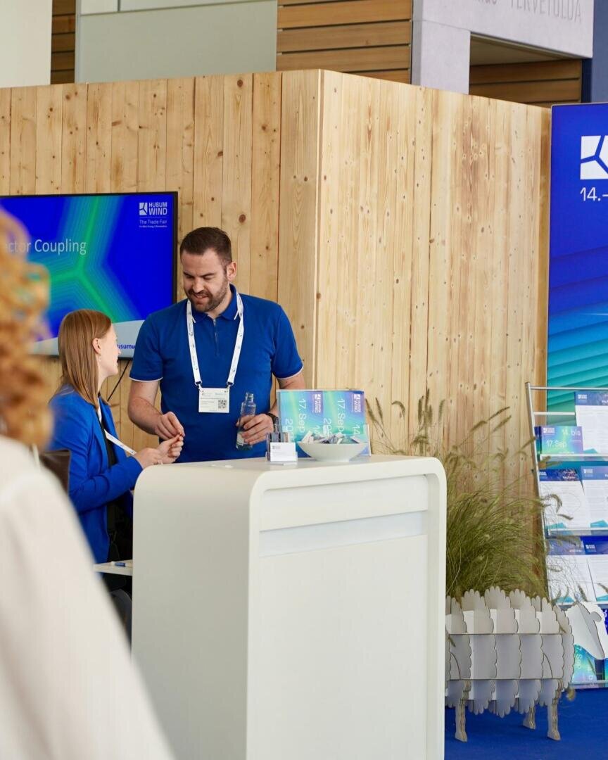 Zwei Personen in blauer Kleidung unterhalten sich an einem weißen Messestand mit Broschüren und einer hölzernen Rückwand. Ein digitaler Bildschirm und Pflanzen sind zu sehen; beide Personen tragen Umhängebänder.
