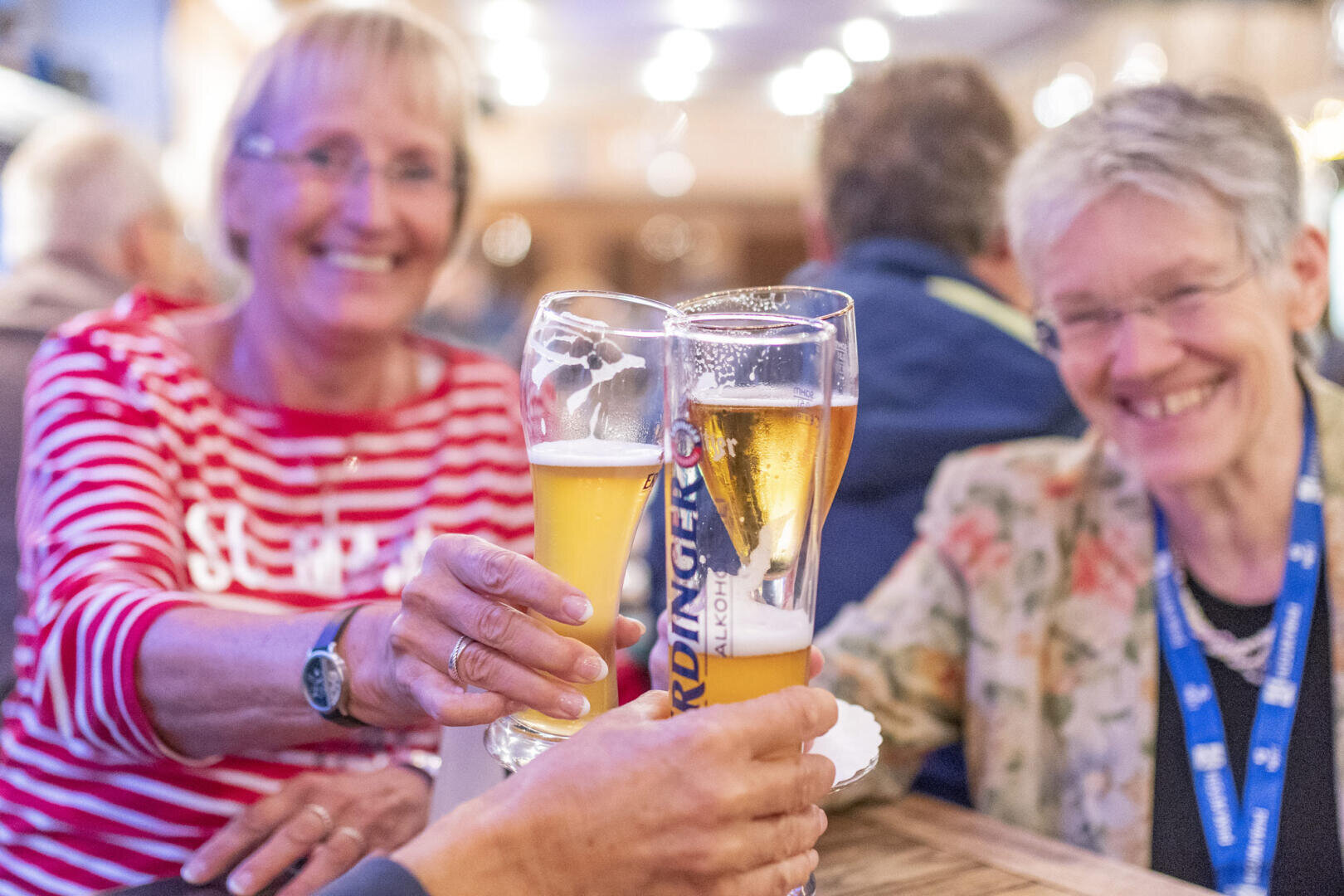 Two smiling elderly women sit at a table, cheerfully clinking large beer glasses, while a lively interior atmosphere prevails in the background.