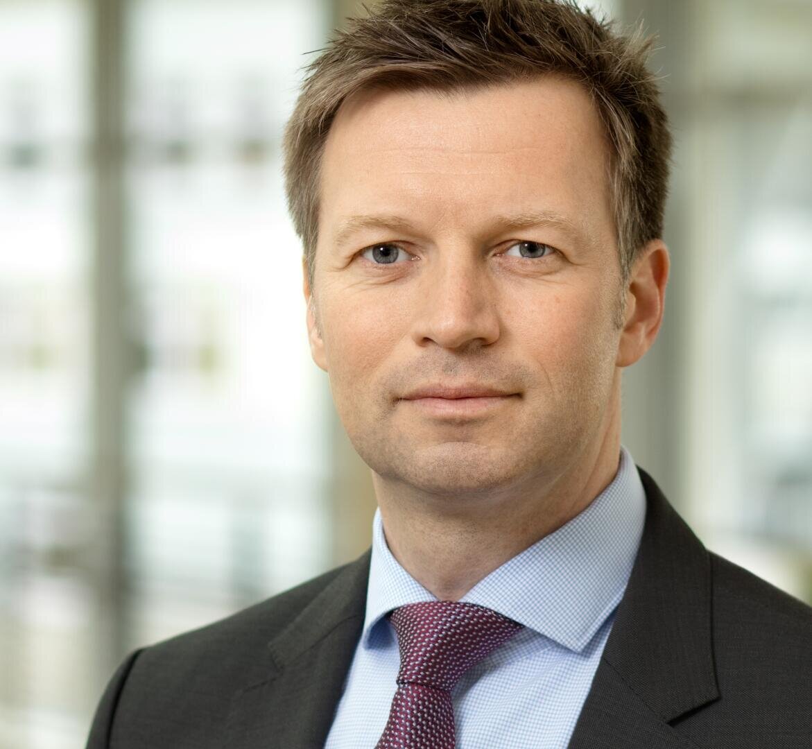 Ein Mann in Anzug und Krawatte mit kurzen braunen Haaren steht in einem Haus und schaut mit neutralem Blick in die Kamera. Der Hintergrund ist unscharf und suggeriert eine Büroumgebung.