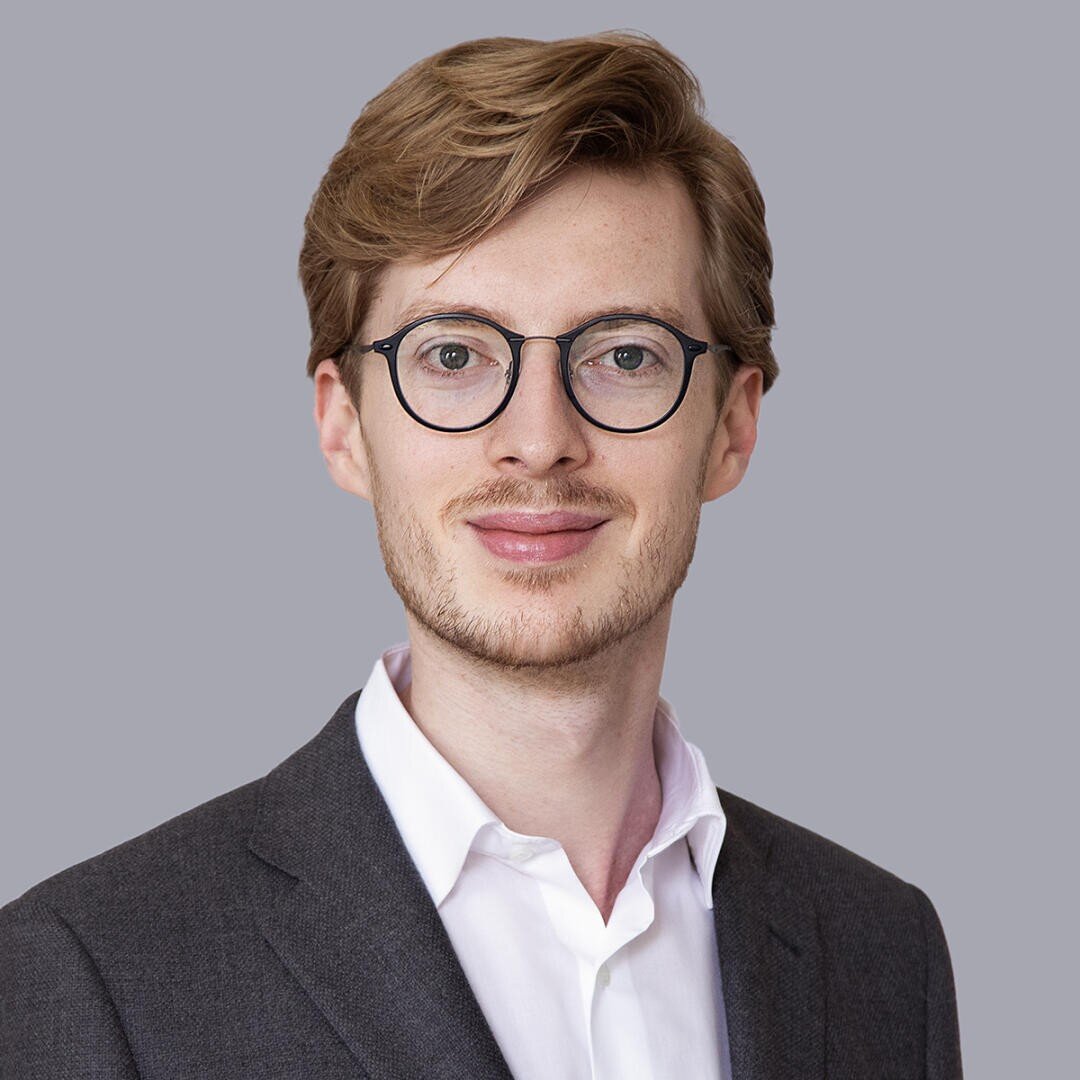 A young man with light brown hair and glasses, wearing a dark suit jacket over a white shirt, smiles softly against a plain light gray background.