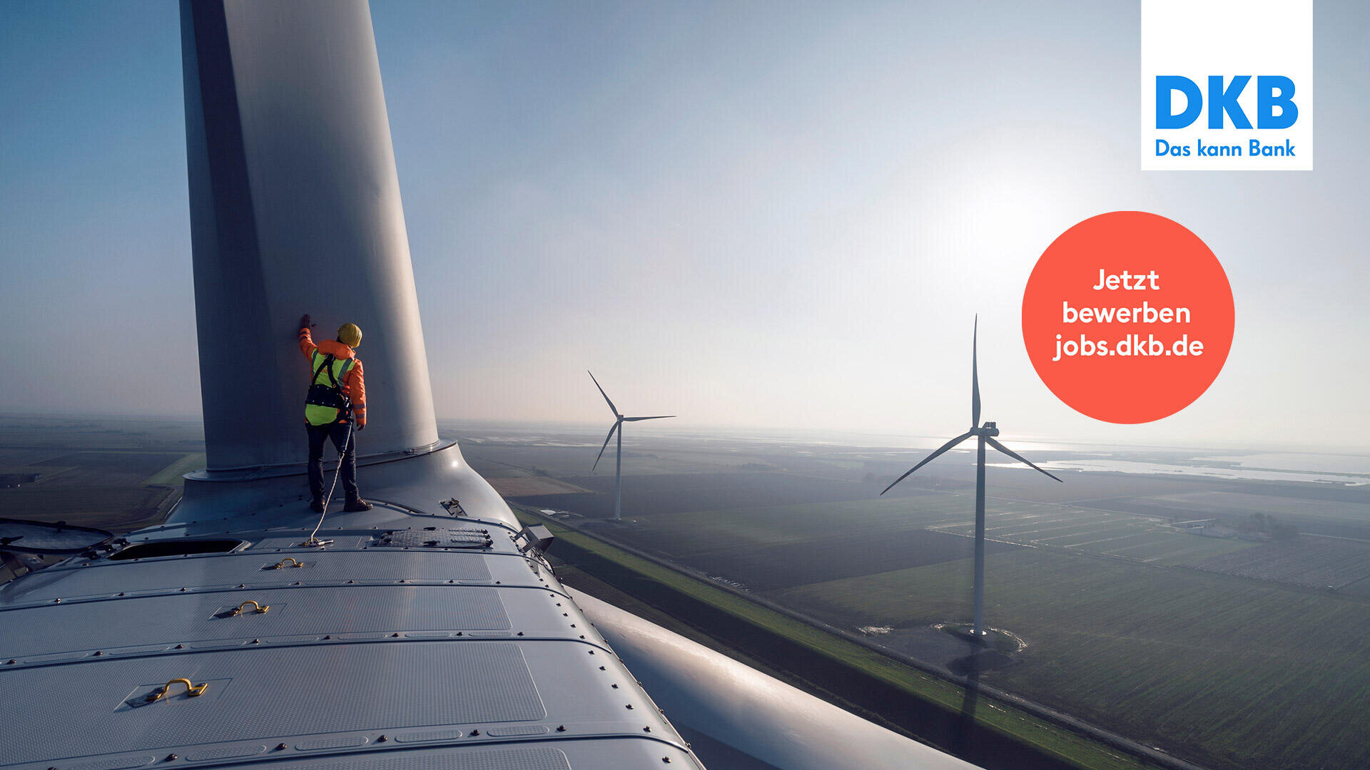 Ein Arbeiter in Schutzkleidung steht auf einer Windkraftanlage und überblickt bei Sonnenaufgang Windparks und Felder. In der oberen rechten Ecke erscheinen das DKB-Logo und ein roter Kreis mit Jetzt bewerben jobs.dkb.de.