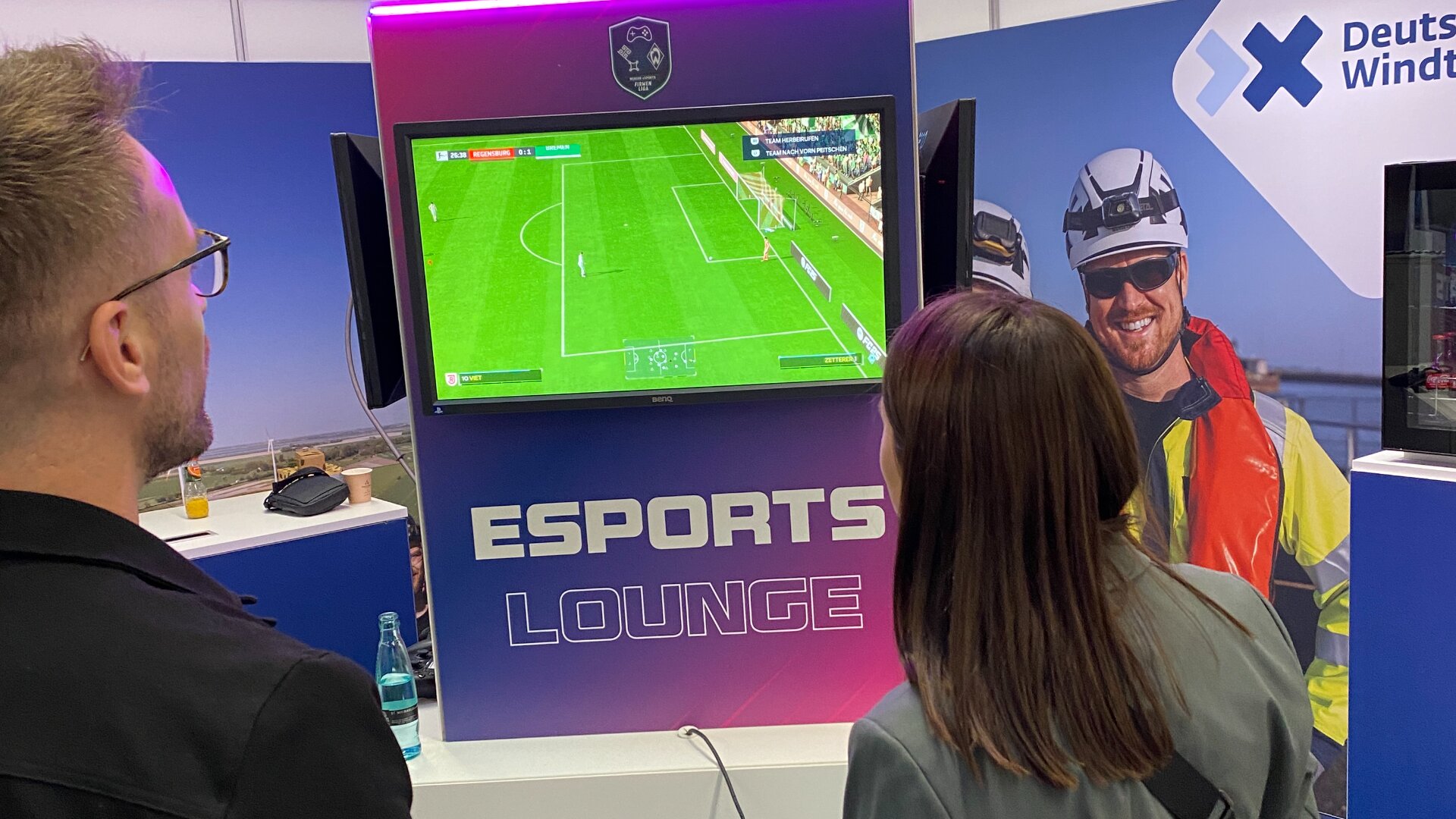 Zwei Personen stehen vor einem Bildschirm und spielen ein Fußballvideospiel an einem Stand der Esports Lounge. Im Hintergrund ist ein großes Poster mit einer lächelnden Person in Schutzausrüstung und dem Logo der Deutschen Windtechnik zu sehen.