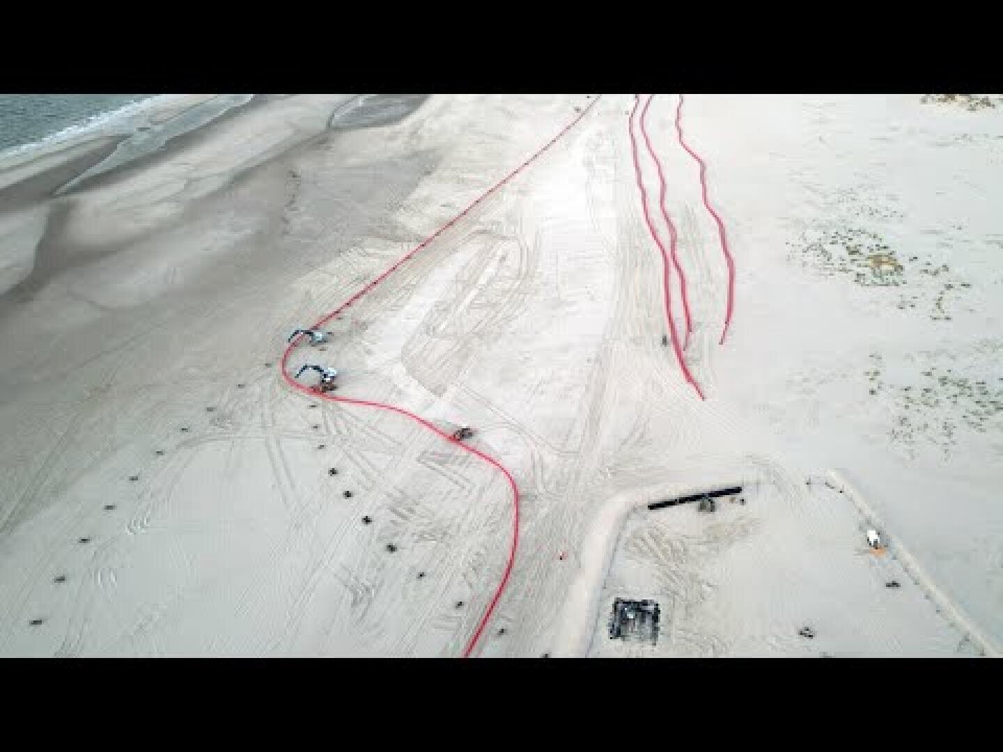 Aerial view of a sandy beach with several long red pipes laid out across the sand, construction vehicles and equipment are visible near the pipes, and tire tracks cover the area.