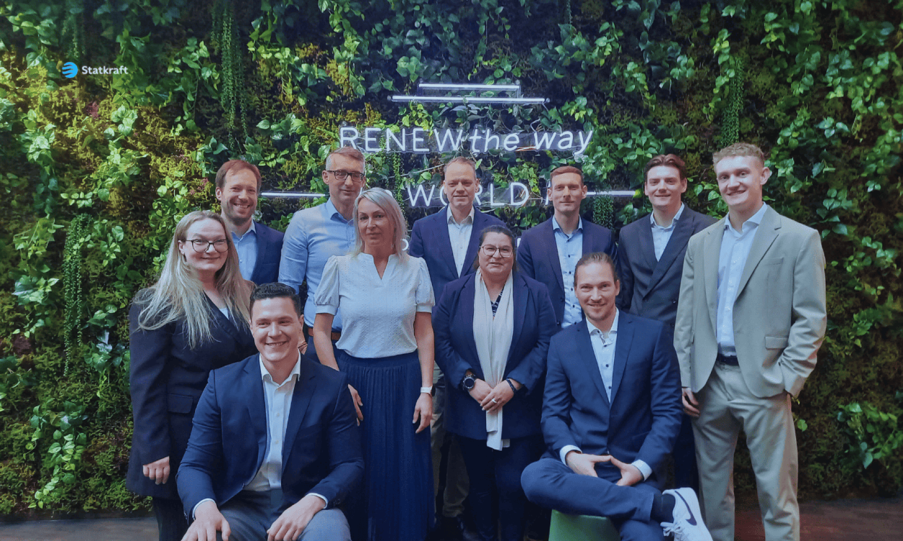 Eine Gruppe von elf Personen in Geschäftskleidung posiert und lächelt vor einer grünen Pflanzenwand mit einer Leuchtreklame, auf der "RENEW the way we change the WORLD" steht. Auf der linken Seite ist das Statkraft-Logo zu sehen.