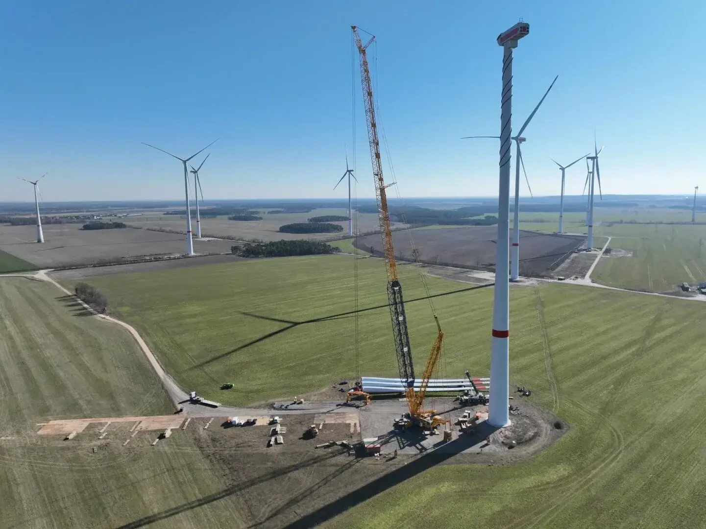 Ein großer Kran montiert eine Windturbine auf einer grünen Wiese, mit Turbinenblättern und Baufahrzeugen auf dem Boden. Im Hintergrund stehen mehrere fertige Windturbinen unter einem klaren blauen Himmel.