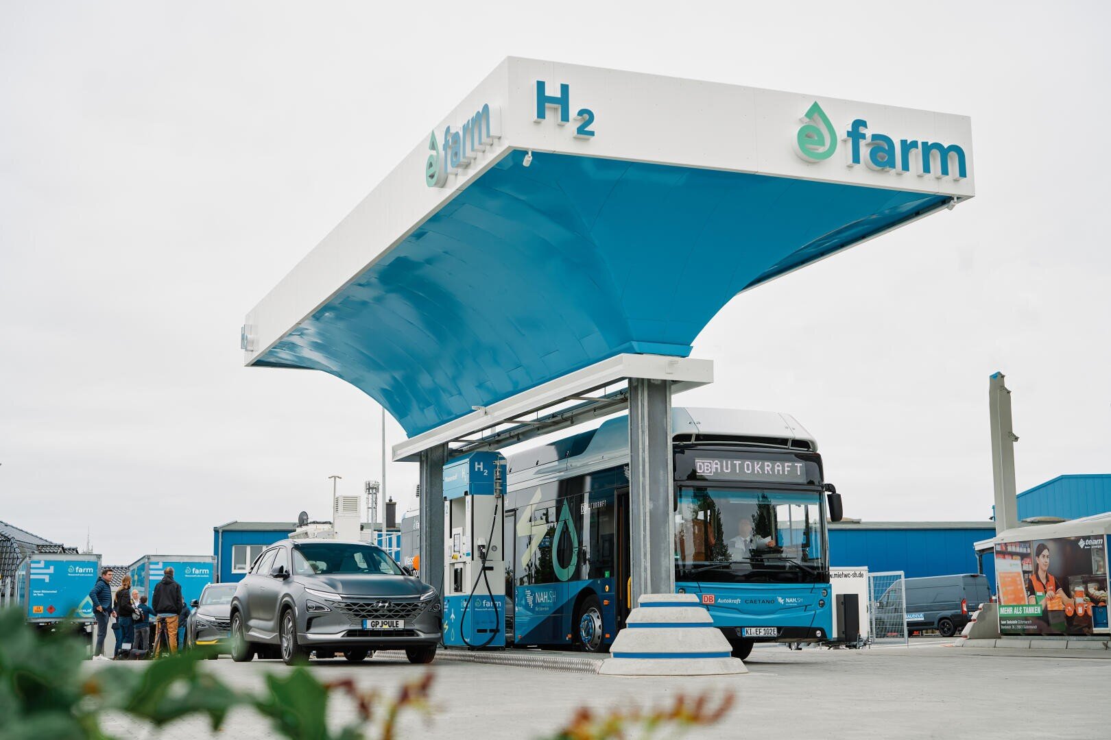 A hydrogen filling station with a large blue and white canopy bearing the words H2 and efarm. A bus and a car are refueling, and several people are standing nearby. The surroundings appear modern, clean, and environmentally friendly.