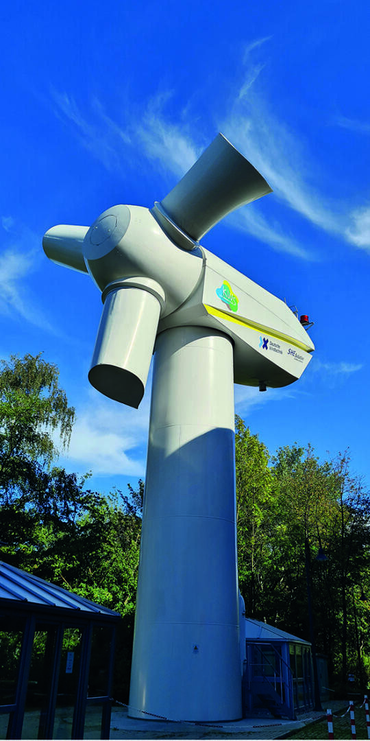 Eine große, weiße Gondel einer Windturbine steht auf einem hohen Sockel im Freien, umgeben von grünen Bäumen unter einem strahlend blauen Himmel mit dünnen Wolken.