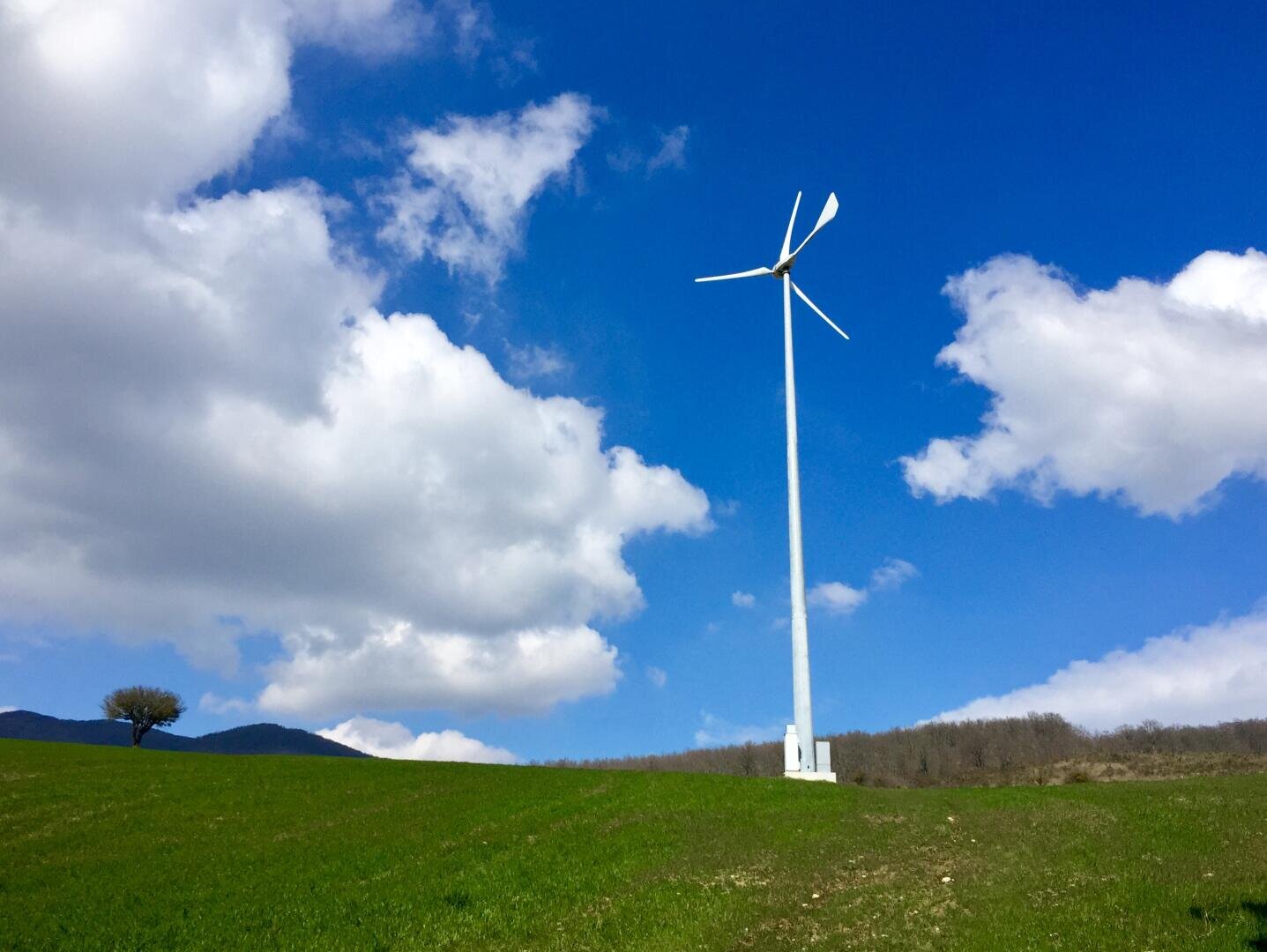 A single wind turbine stands on a grassy hill under a bright blue sky with scattered white clouds. There is a solitary tree on the left and a line of trees in the background.