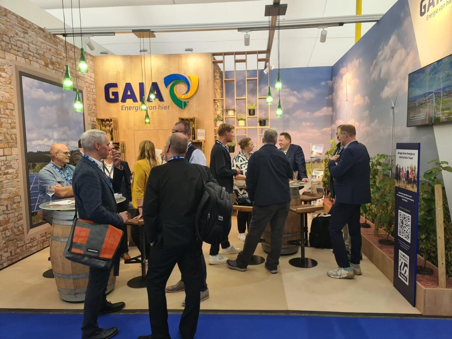 A group of people gathers and converses at a trade show booth with a GAIA sign, featuring wood decor, a plant, and informational displays. Some hold bags and wear name badges.
