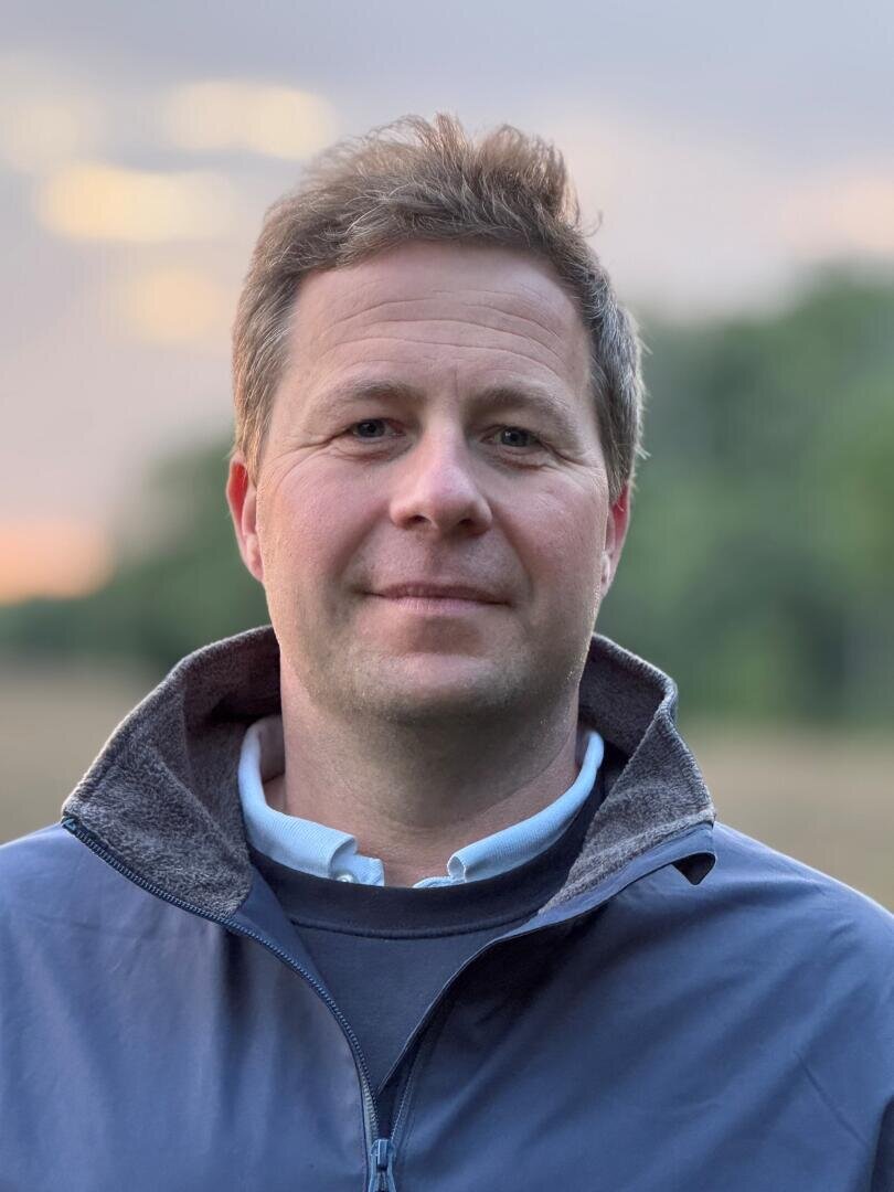 A man with short brown hair wearing a blue jacket and white collared shirt stands outdoors, with a blurred background of greenery and a soft sunset sky.