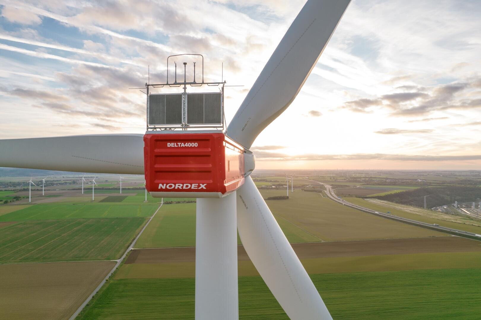 Nahaufnahme einer großen Nordex Delta4000-Windturbine in einer ländlichen Landschaft bei Sonnenuntergang, wobei die Flügel und die rote Gondel sichtbar sind. Im Hintergrund sind Felder und weitere Windkraftanlagen zu sehen.