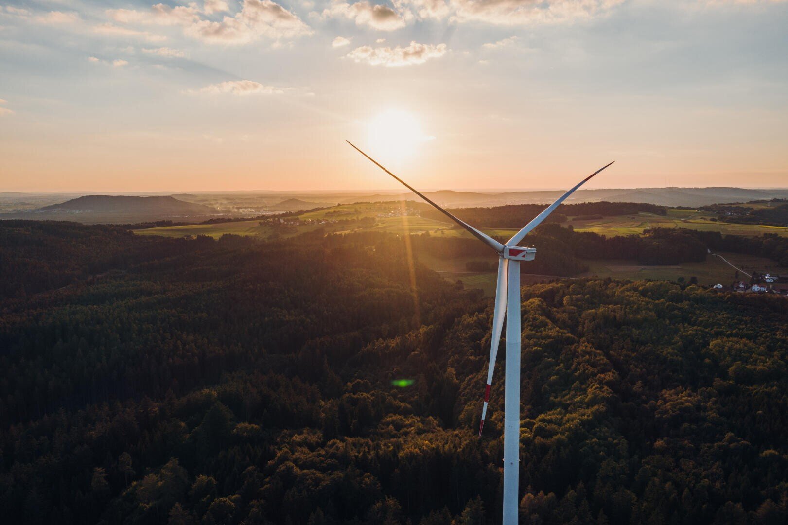 A wind turbine stands tall over a forested landscape at sunset, with the sun low in the sky and fields, trees, and a few scattered houses in the distance.