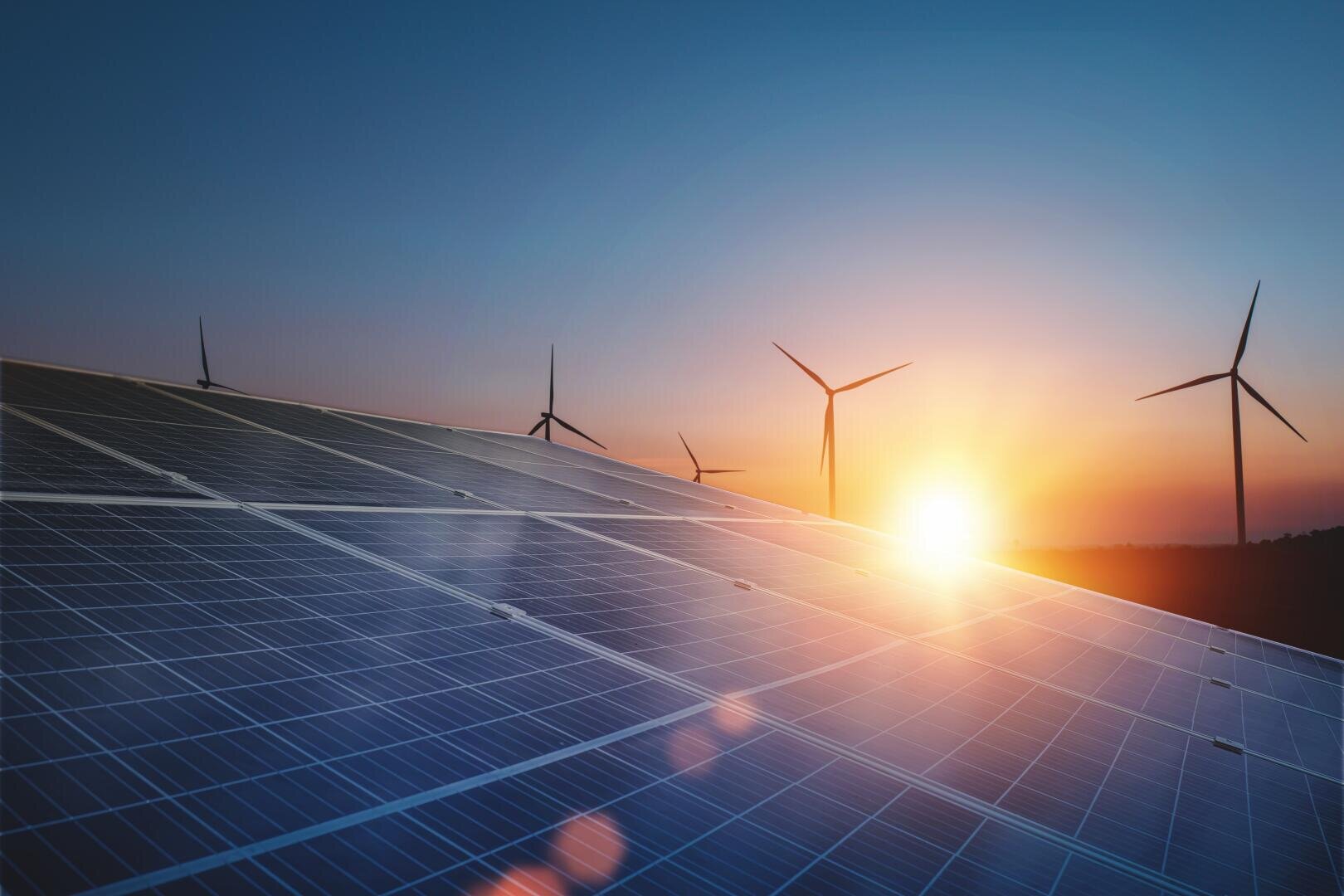 Reihen von Solarmodulen und Windturbinen werden bei Sonnenaufgang oder Sonnenuntergang vor einem klaren Himmel aufgestellt, wobei die erneuerbaren Energiequellen durch die am Horizont leuchtende Sonne hervorgehoben werden.