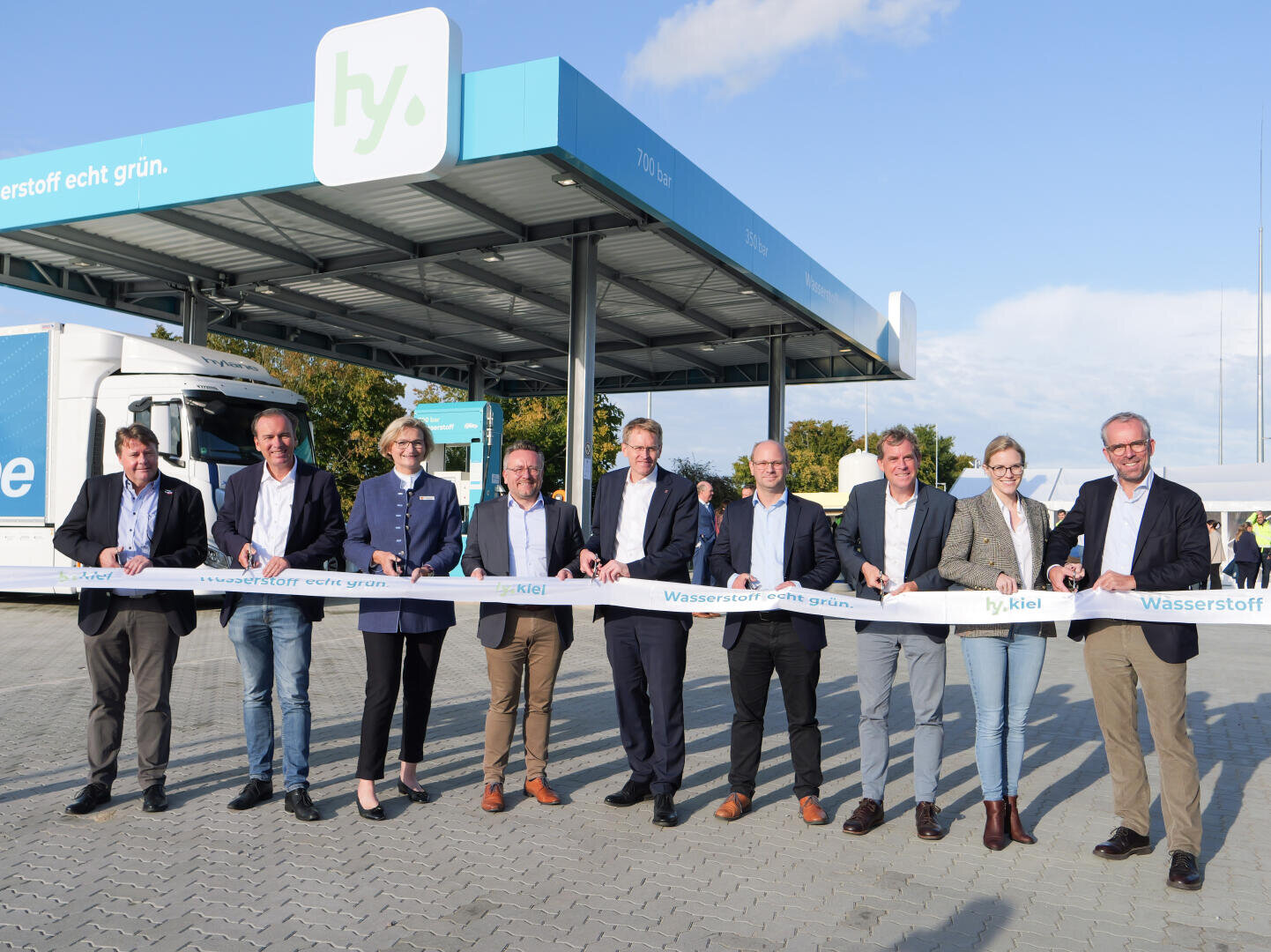 Neun Personen in Geschäftskleidung stehen vor einer Wasserstofftankstelle und halten ein Band zur feierlichen Eröffnung. Ein blau-weißer Lastwagen ist in der Nähe geparkt, und auf einem Schild über der Tankstelle steht "hy." in grün und weiß.