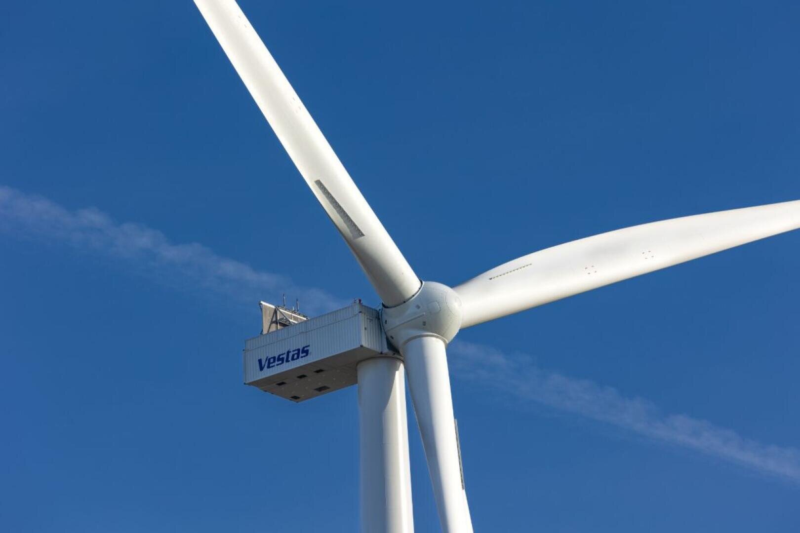 Nahaufnahme einer weißen Vestas-Windturbine vor strahlend blauem Himmel. Zu sehen sind die Rotorblätter und die Gondel der Turbine, auf der das Vestas-Logo zu sehen ist.