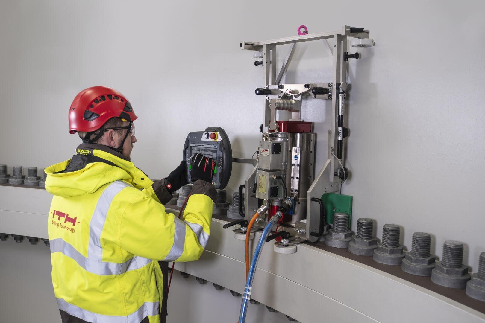 A worker in a yellow high-visibility jacket and red helmet operates a control panel connected to a large industrial machine mounted on a wall with rows of heavy bolts.