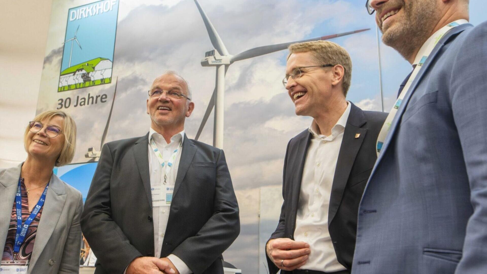 Four adults in business attire stand smiling in front of a display with a wind turbine image and the text DIRKSHOF 30 Jahre against a cloudy sky background.