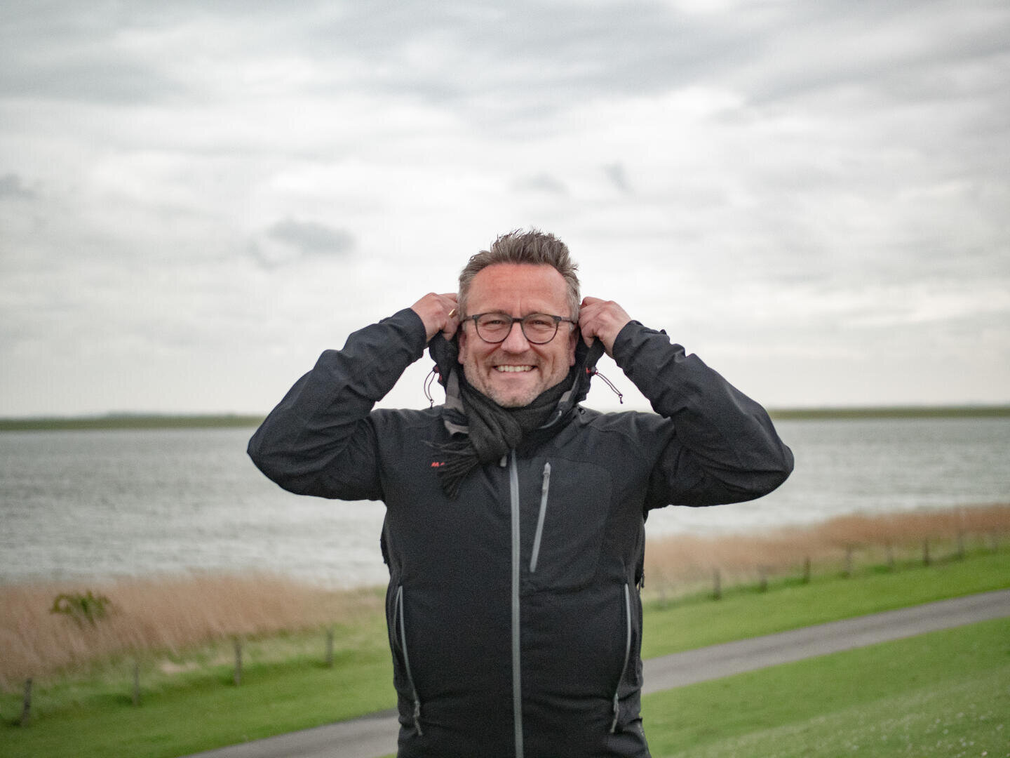 A smiling man wearing glasses and a black jacket stands outdoors near a body of water, holding his hood with both hands. The background shows grass, a path, and cloudy sky.