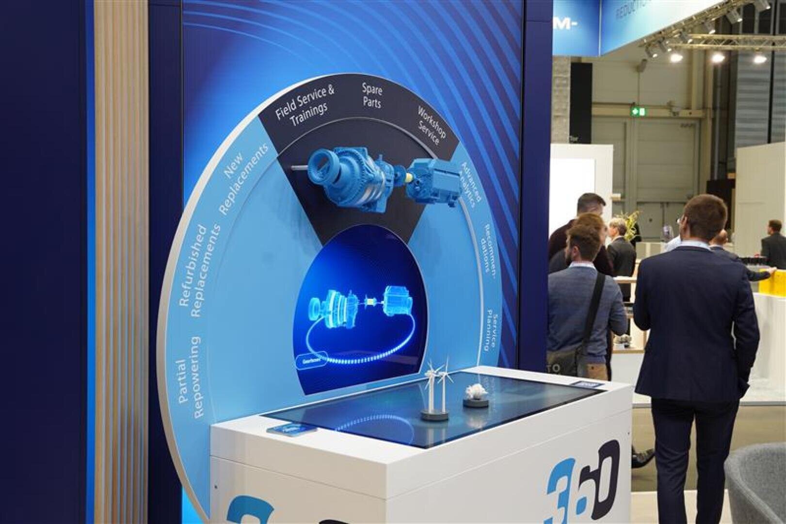 Auf einem Messestand ist ein blaues Kreisdiagramm über Außendienst und Ersatzteile zu sehen, darunter 3D-Modelle von Industriekomponenten und ein kleiner Tisch. Mehrere Personen stehen in der Nähe und unterhalten sich.