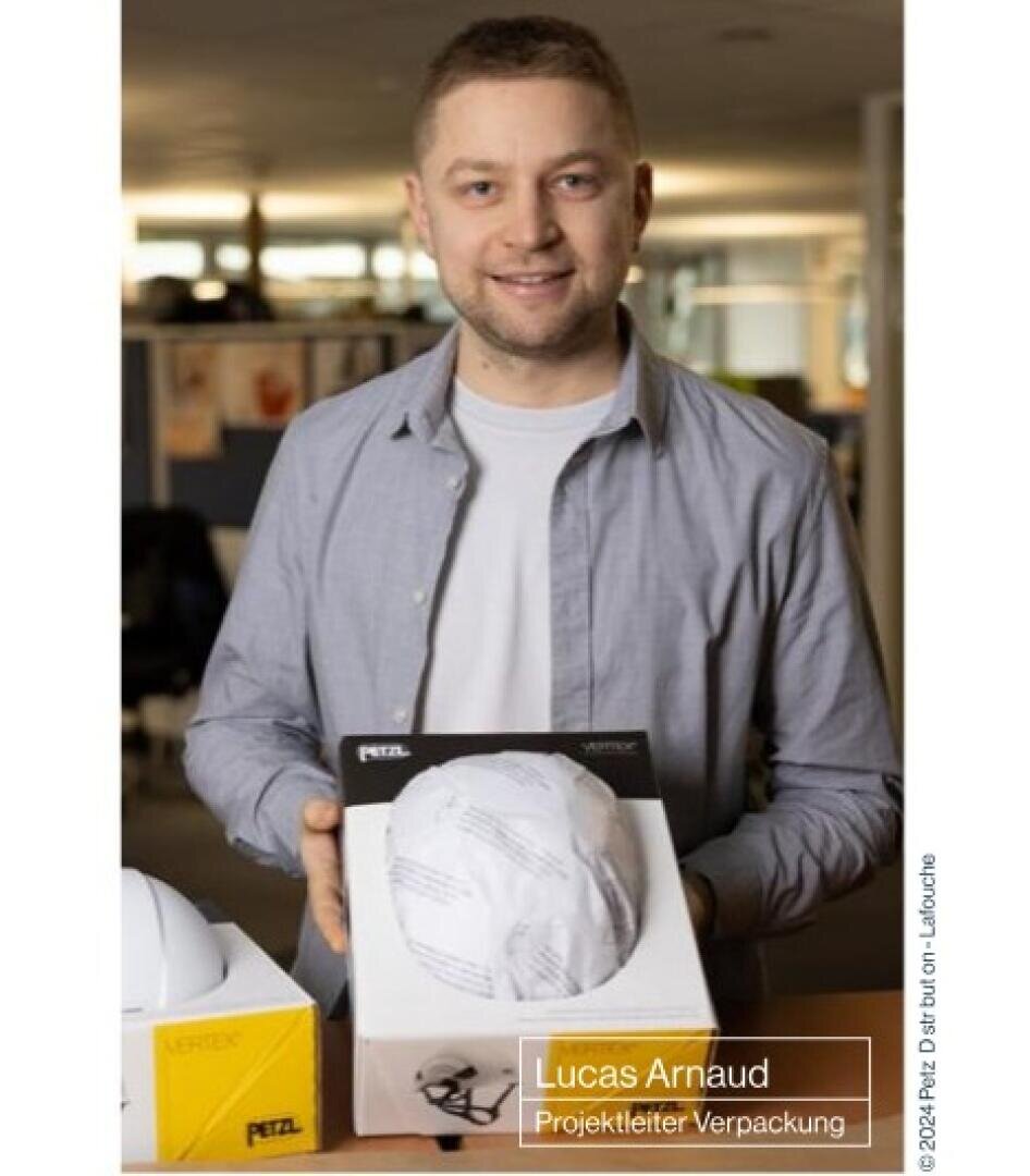 Ein Mann lächelt, während er einen verpackten Helm an einem Bürotisch präsentiert. Der Text auf dem Bild lautet: Lucas Arnaud, Projektleiter Verpackung. Auf dem Schreibtisch sind auch andere Schachteln und Helme zu sehen.
